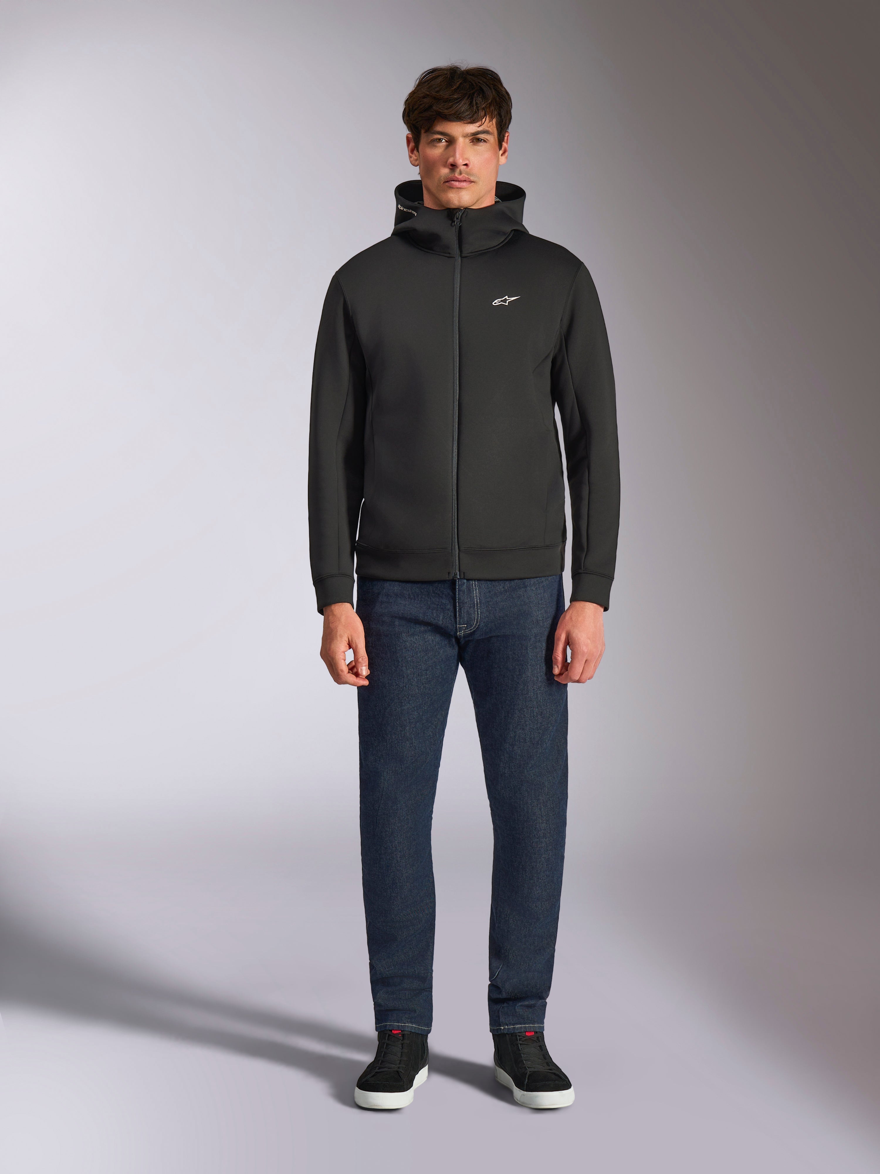 Modell trägt den Alpinestars Cypher Hoodie, schwarzer Fleece-Hoodie mit Front-Reißverschluss, hochgeschlossene Kapuze mit kleinem weißen Astars-Logo auf der Brust, kombiniert mit dunkler Denim-Jeans und schwarzen Sneakern, lässige Motorsport-inspirierte Streetwear