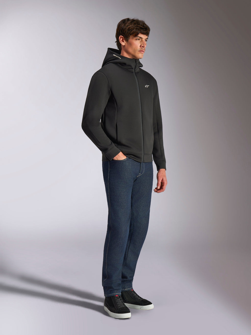 Alpinestars Cypher Hoodie, lässige Streetwear, schwarzer Kapuzenpullover mit Reißverschluss, männliches Model in dunkelblauen Jeans und schwarzen Sneakern, hochgeschlossene Kapuze mit Alpinestars-Wortlogo, kleines weißes Astars-Logo auf der Brust, eleganter vom Motorsport inspirierter Lifestyle-Look