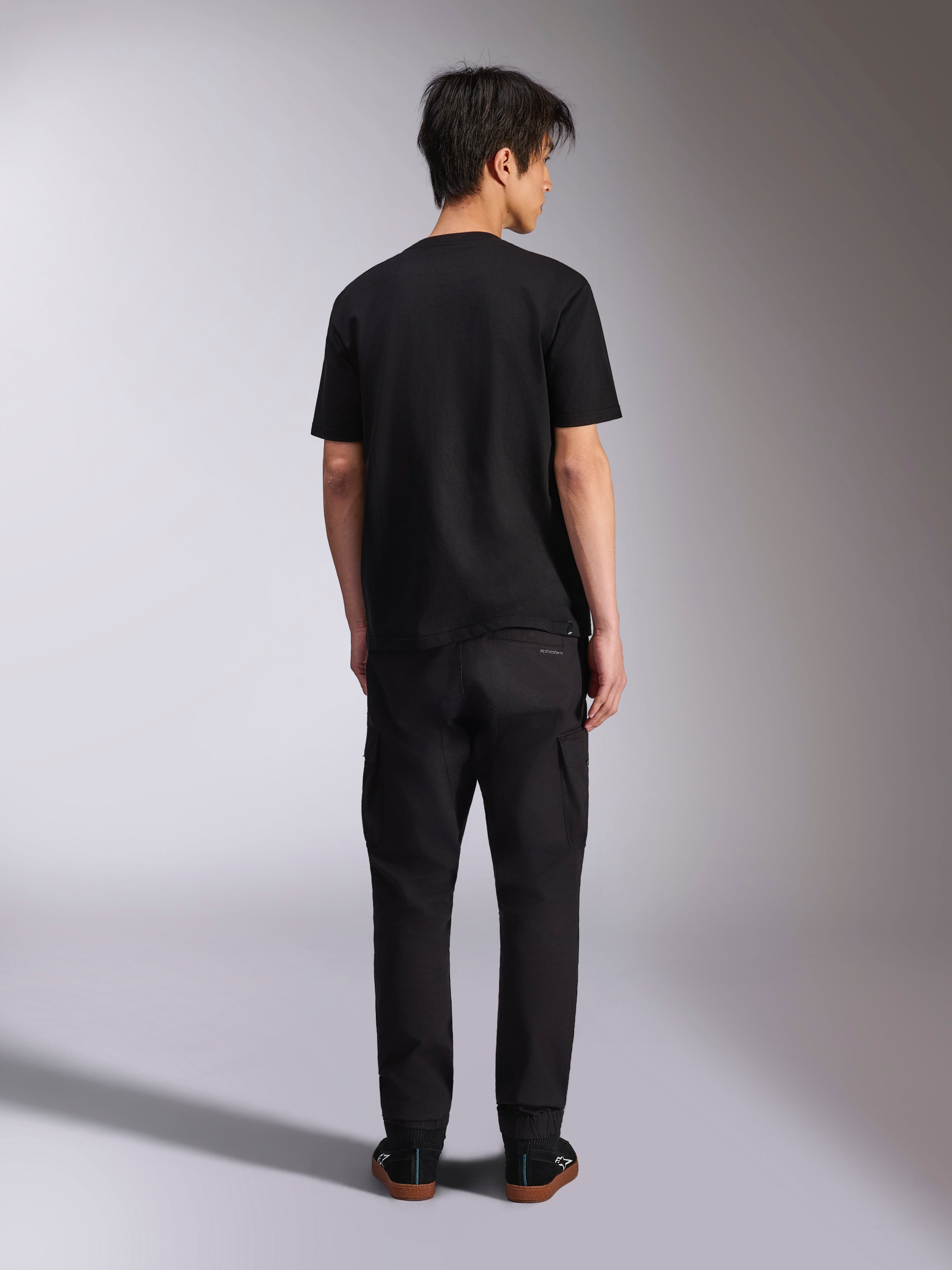 Alpinestars Noka CSF Tee, kurzärmeliges T-Shirt, Schwarz, Rückansicht eines Models, das das T-Shirt mit schwarzer Hose und Sneakern trägt, lässiger Motorsport-Streetwear-Stil