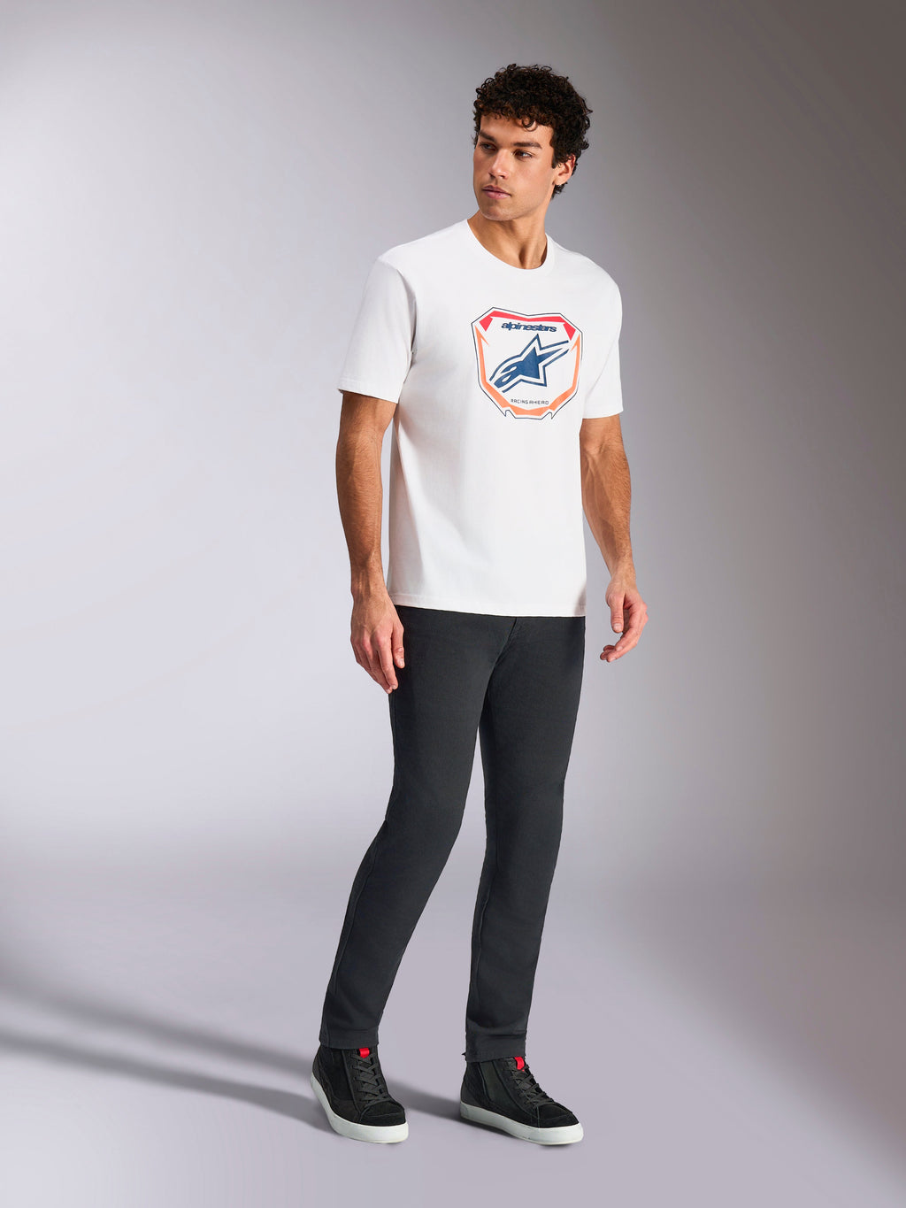Alpinestars Plate CSF Tee, Kurzarm-T-Shirt, Weiß, mit einer Astars MX Plattengrafik in Blau und Orange auf der Vorderseite, lässige motorsport-inspirierte Streetwear getragen mit dunkler Hose und High-Top-Sneakern
