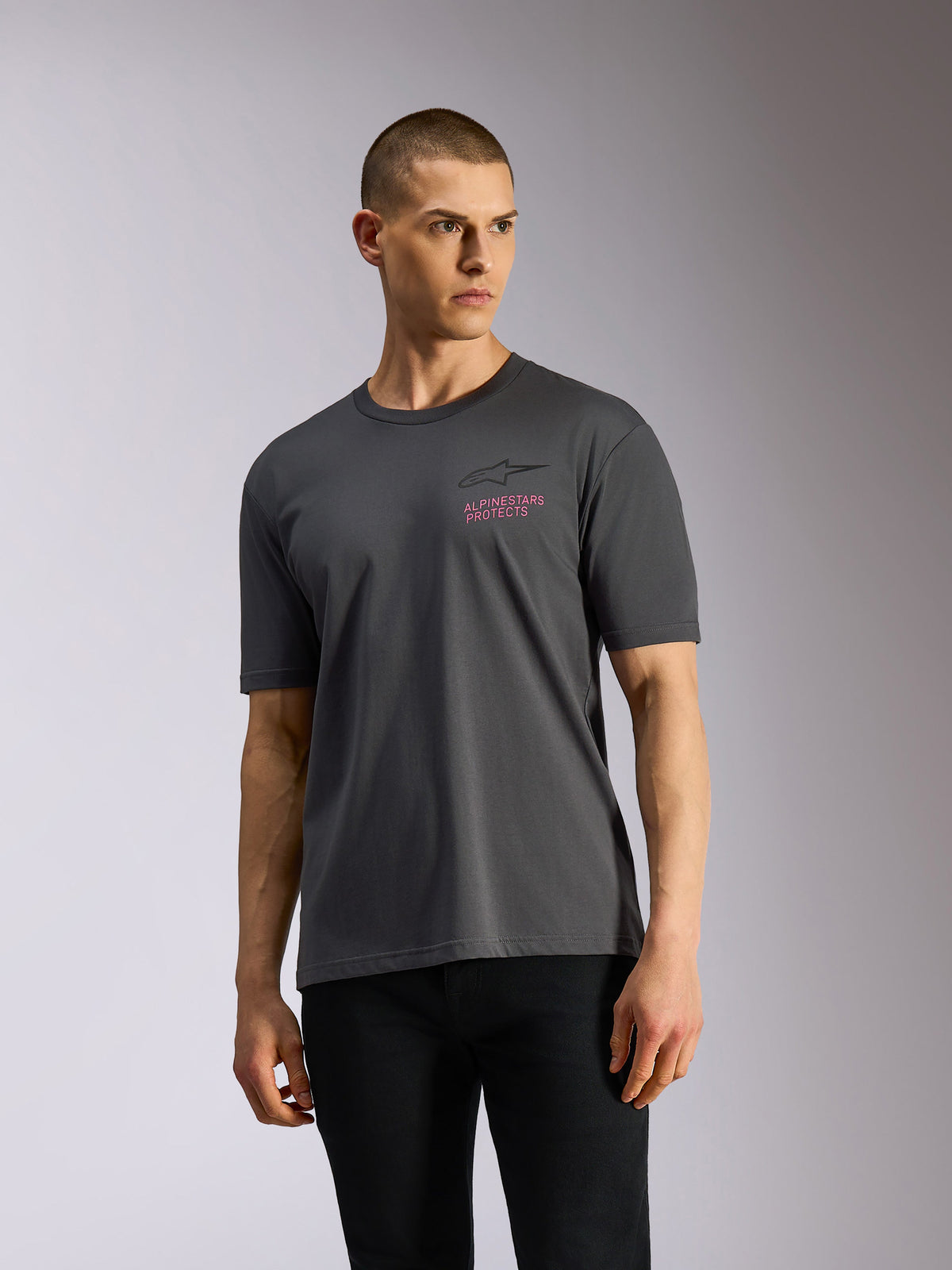 Alpinestars Pop Helmet CSF Tee, kurzärmeliges T-Shirt, Anthrazitgrau, von einem Model getragen, mit schwarzem Astars-Logo und pinkfarbenem „ALPINESTARS PROTECTS“-Schriftzug auf der Brust, lässige Motorsport-Streetwear