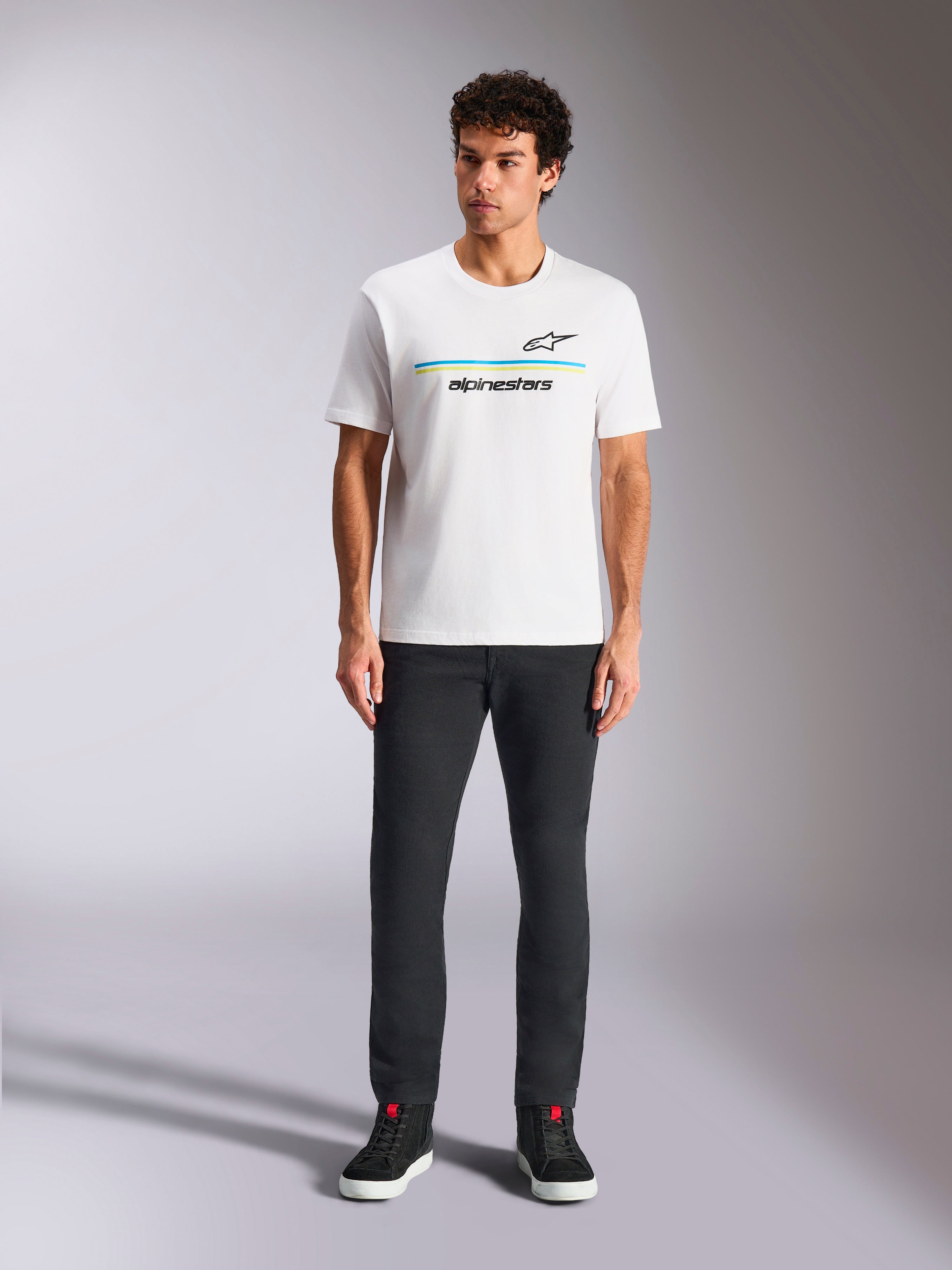 Alpinestars Lanes CSF Tee, Kurzarm-T-Shirt, Weiß, blau-gelbe Streifen-Logo-Grafik, getragen von Model mit schwarzer Hose und Sneakern, lässige Motorsport-Streetwear