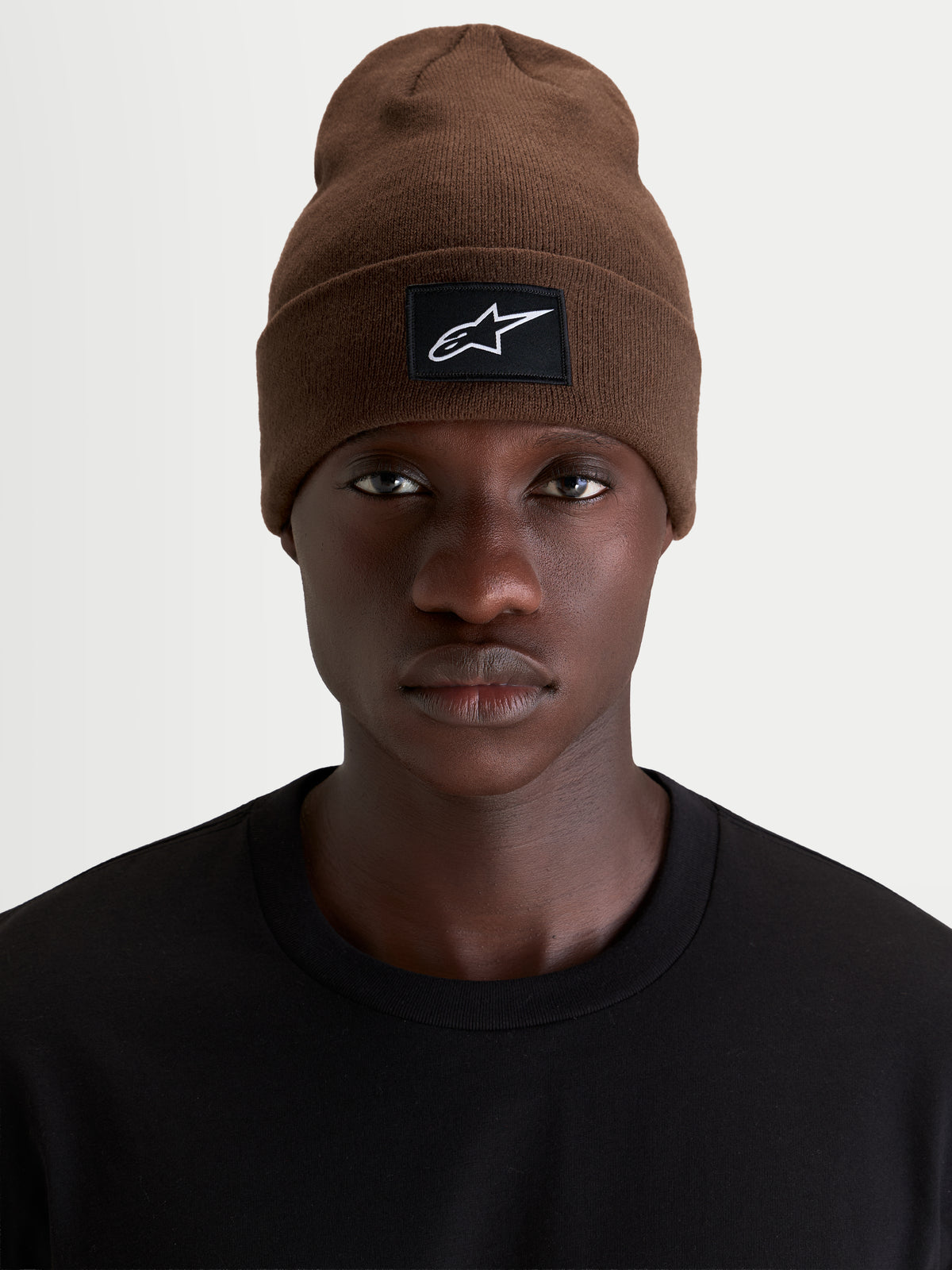 Alpinestars Augment Cuff Mütze, Sportswear-Mütze in Braun mit gewebtem Alpinestars Logo-Patch am Umschlag, getragen von einem Model mit schwarzem T-Shirt