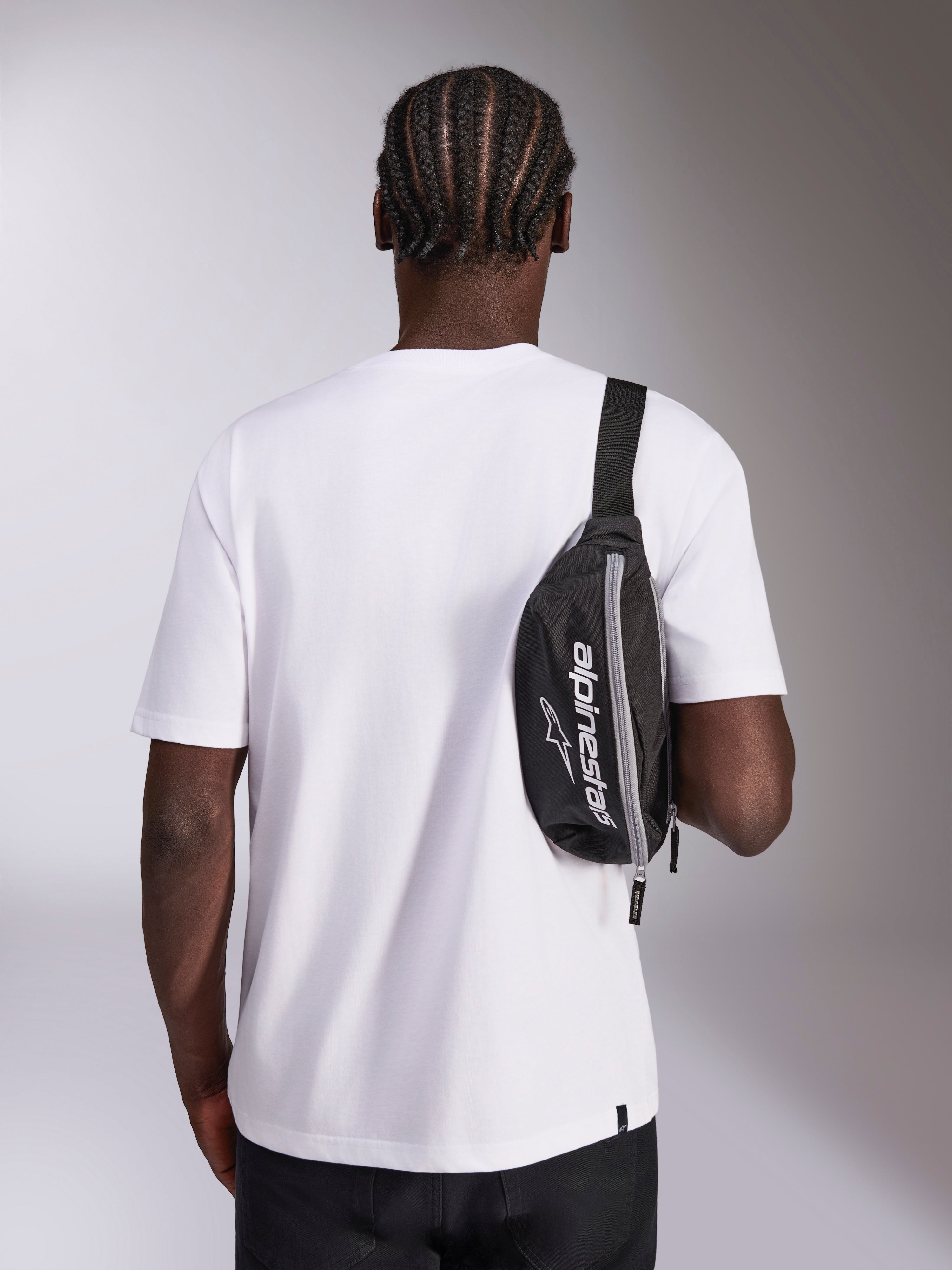 Alpinestars Only Linear Hüfttasche, schwarz mit weißem Alpinestars-Logo, als Crossbody-Tasche über einem weißen T-Shirt getragen, lässiges, vom Motorsport inspiriertes Streetwear-Accessoire