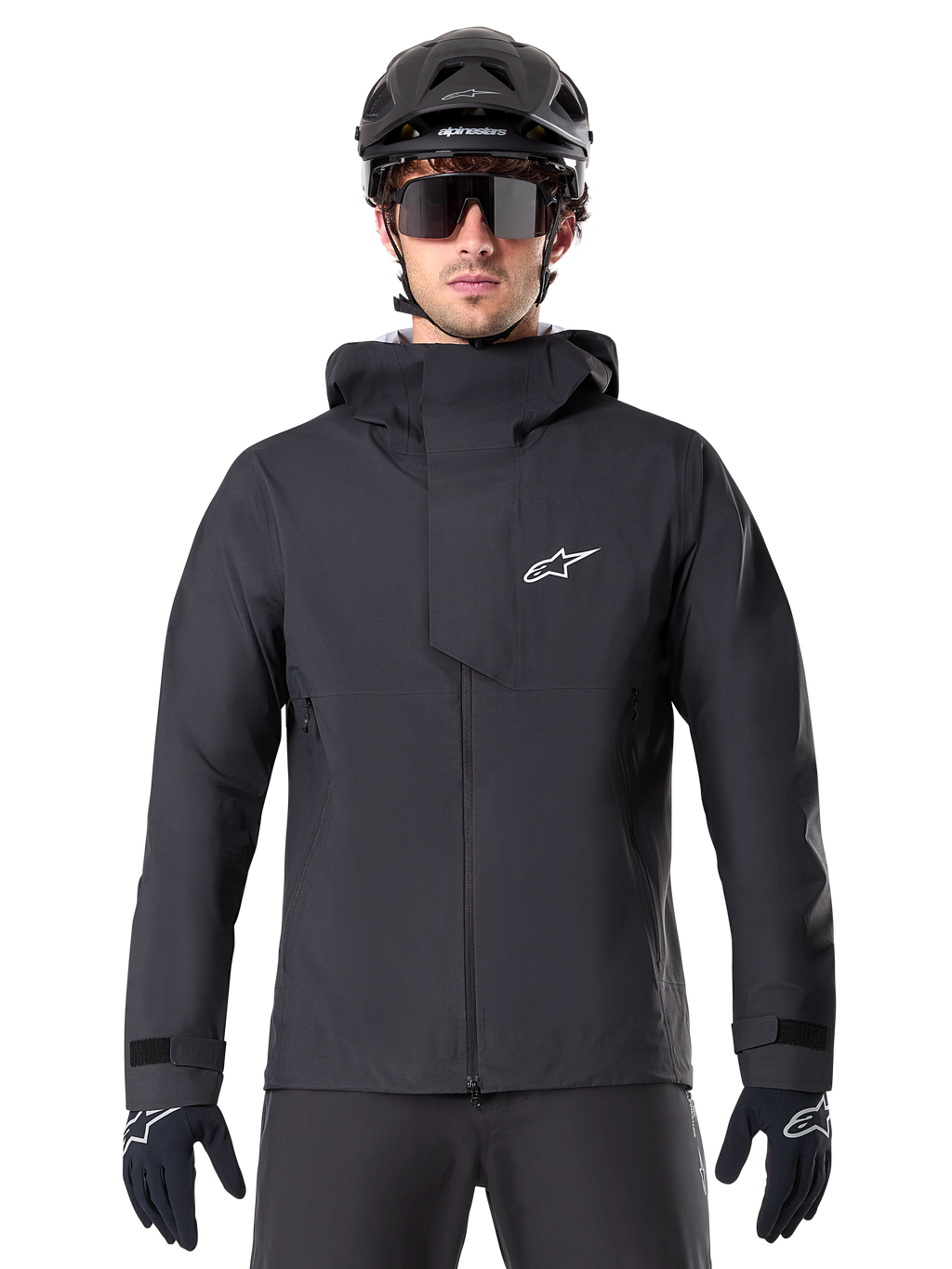 Alpinestars A-Dura Elite Wasserdichte Jacke, Schwarz, Mountainbike-Jacke, Design mit hohem Kragen und Kapuze, Frontreißverschluss mit schützender Brustklappe und weißem Logo, getragen mit schwarzem Helm und Fahrradhandschuhen, technischer Wetterschutz für Trail-Fahrten