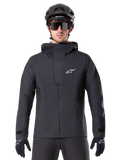 Alpinestars A-Dura Elite Wasserdichte Jacke, Schwarz, Mountainbike-Jacke, Design mit hohem Kragen und Kapuze, Frontreißverschluss mit schützender Brustklappe und weißem Logo, getragen mit schwarzem Helm und Fahrradhandschuhen, technischer Wetterschutz für Trail-Fahrten