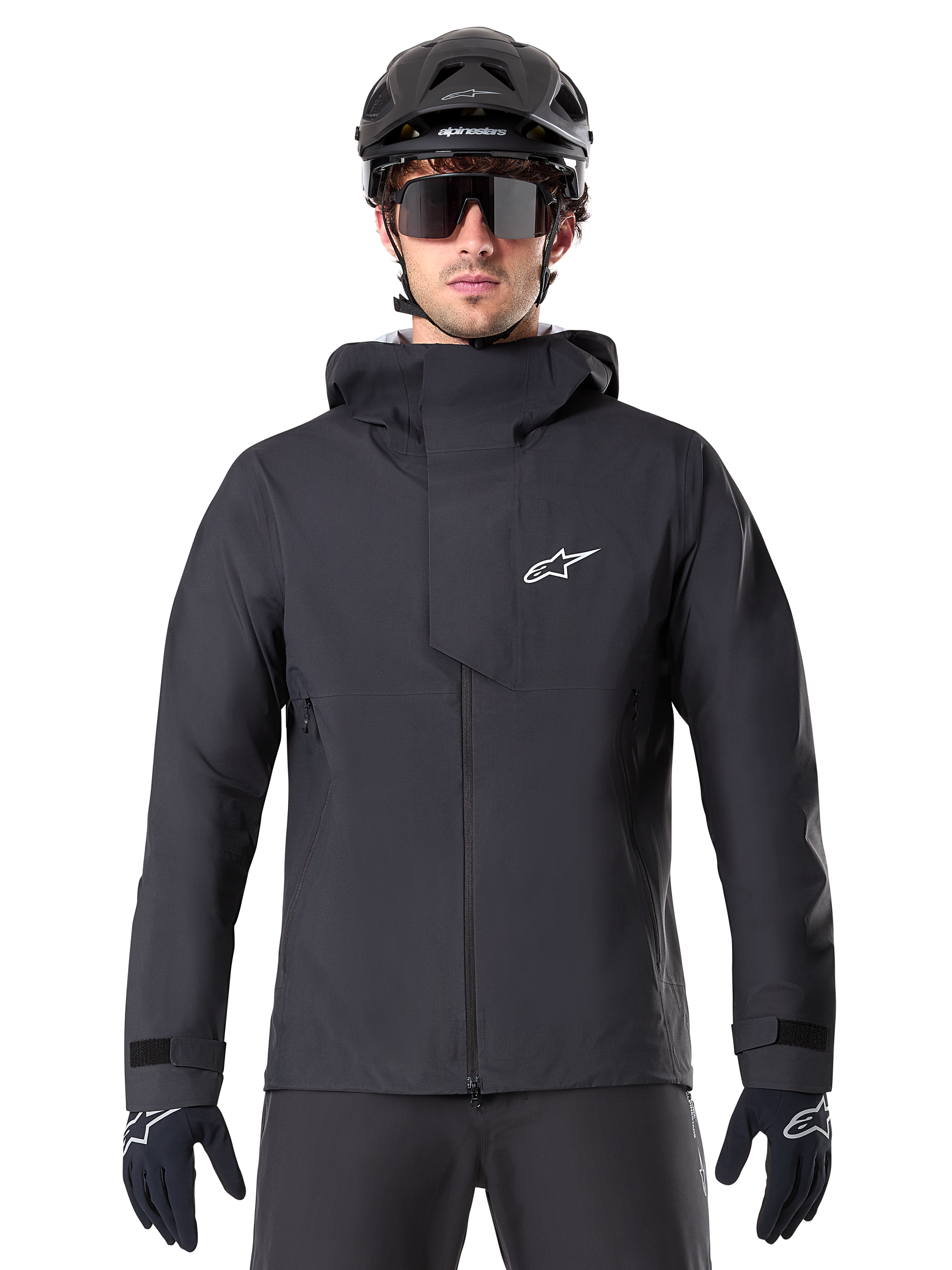 Alpinestars A-Dura Elite Wasserdichte Jacke, Schwarz, Mountainbike-Jacke, Design mit hohem Kragen und Kapuze, Frontreißverschluss mit schützender Brustklappe und weißem Logo, getragen mit schwarzem Helm und Fahrradhandschuhen, technischer Wetterschutz für Trail-Fahrten