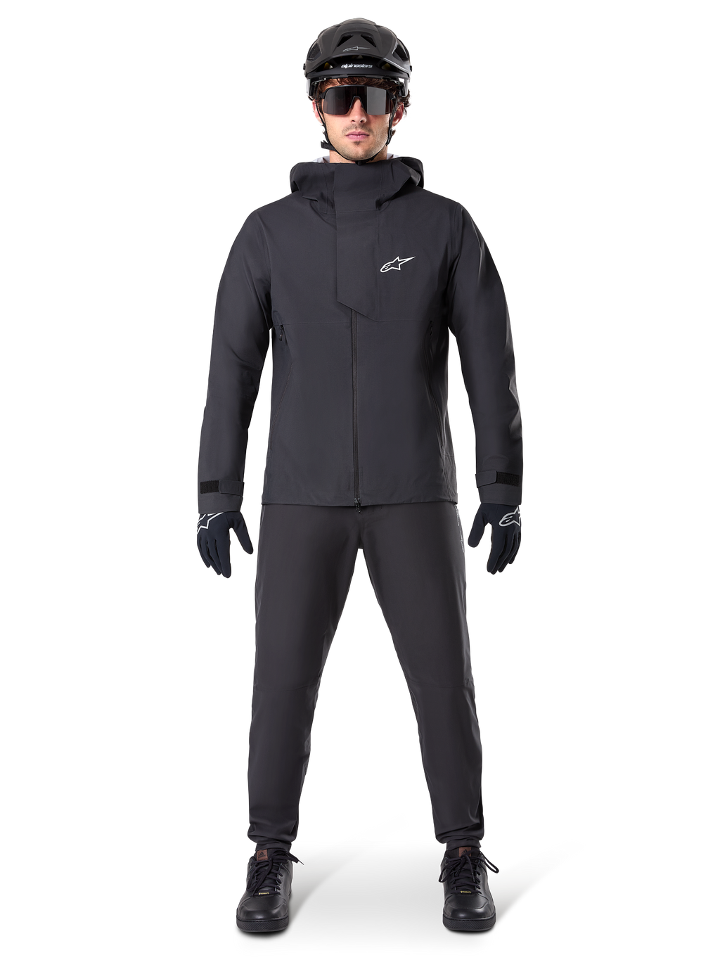 Alpinestars A-Dura Elite Wasserdichte Jacke, Schwarz, getragen mit passender Fahrradhose und Handschuhen, schwarzer Helm und Sonnenbrille, mit weißem Astars-Logo auf der Brust, leistungsstarkes technisches Design für Mountainbiking