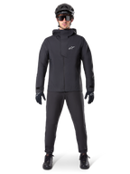 Alpinestars A-Dura Elite Wasserdichte Jacke, Schwarz, getragen mit passender Fahrradhose und Handschuhen, schwarzer Helm und Sonnenbrille, mit weißem Astars-Logo auf der Brust, leistungsstarkes technisches Design für Mountainbiking