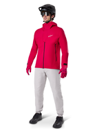 Male cyclist wearing the Alpinestars A-Dura Elite Waterproof Jacket in Red Fluo, paired with light grey technical pants, red gloves, black MTB helmet with goggles, and black shoes, complete high-performance outfit for mountain biking