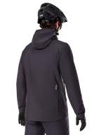 Alpinestars A-Dura Thermojacke, Schwarz, Rückansicht, getragen mit einem schwarzen Mountainbike-Helm und Handschuhen, kleines weißes Astars-Logo auf dem unteren Rücken, leichte isolierte Radjacke
