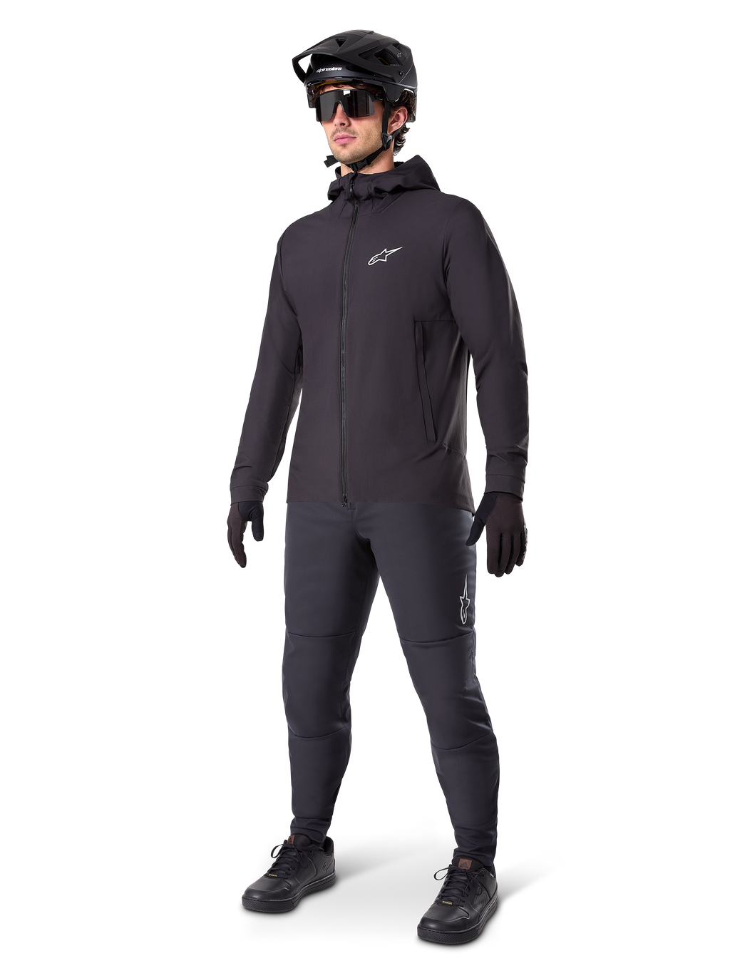 Alpinestars A-Dura Thermojacke in Schwarz, getragen von einem männlichen Fahrer mit kompletter Mountainbike-Ausrüstung inklusive schwarzem Helm, Sonnenbrille und Radhose, leichte isolierte Kapuzenjacke für Radsport-Performance bei kaltem Wetter.