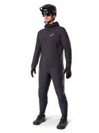 Alpinestars A-Dura Thermojacke in Schwarz, getragen von einem männlichen Fahrer mit kompletter Mountainbike-Ausrüstung inklusive schwarzem Helm, Sonnenbrille und Radhose, leichte isolierte Kapuzenjacke für Radsport-Performance bei kaltem Wetter.