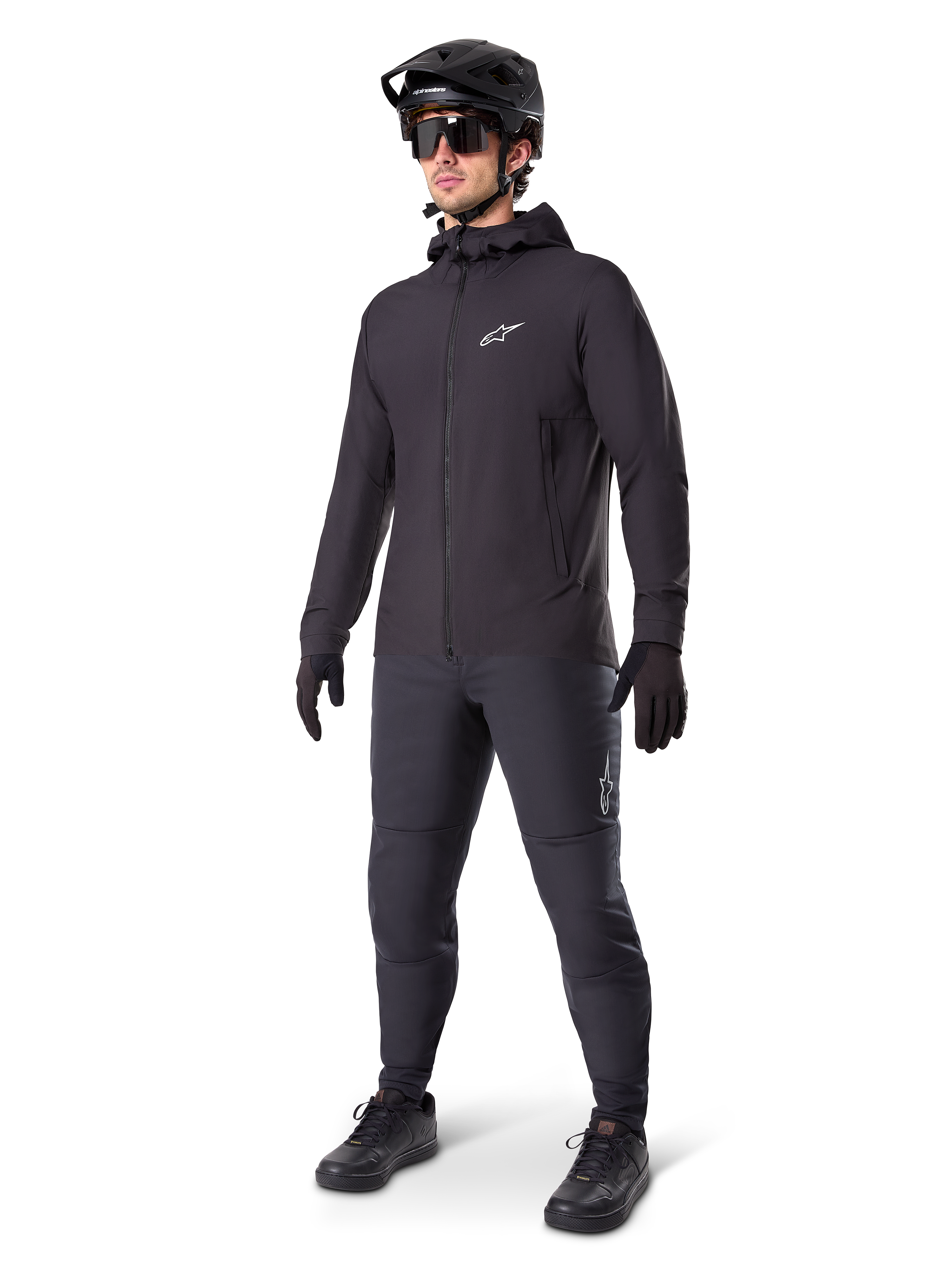 Alpinestars A-Dura Thermojacke in Schwarz, getragen von einem männlichen Fahrer mit kompletter Mountainbike-Ausrüstung inklusive schwarzem Helm, Sonnenbrille und Radhose, leichte isolierte Kapuzenjacke für Radsport-Performance bei kaltem Wetter.