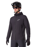 Alpinestars A-Dura Thermal Jacket, schwarze Fahrradjacke, Design mit Kapuze und Frontreißverschluss, weißes Stern-Logo auf der Brust, getragen mit schwarzem Mountainbike-Helm und Handschuhen, sportliche Funktionsbekleidung für Trailfahrten bei kaltem Wetter