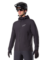 Alpinestars A-Dura Thermal Jacket, schwarze Fahrradjacke, Design mit Kapuze und Frontreißverschluss, weißes Stern-Logo auf der Brust, getragen mit schwarzem Mountainbike-Helm und Handschuhen, sportliche Funktionsbekleidung für Trailfahrten bei kaltem Wetter