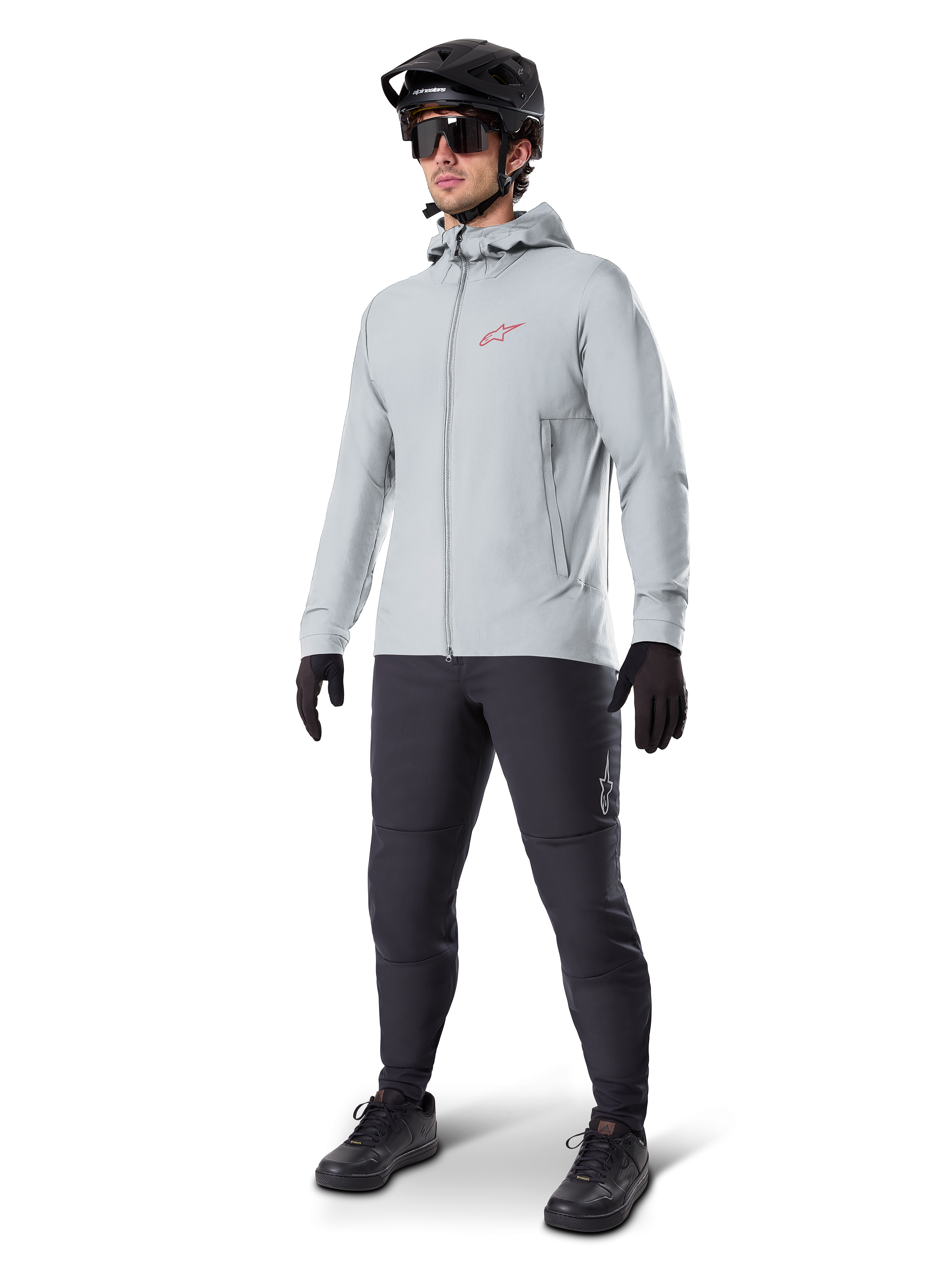 Alpinestars A-Dura Thermal Jacket in Titanium, light gray hooded cycling jacket with a red Alpinestars logo on the chest, worn by a man with a black MTB helmet, sunglasses, dark gray pants, and black gloves, professional mountain bike gear