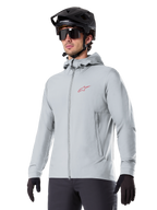 Alpinestars A-Dura Thermal Jacket, light gray insulated cycling jacket, hooded design with a small red logo, worn with a black helmet and gloves, technical mountain bike apparel for cold-weather riding