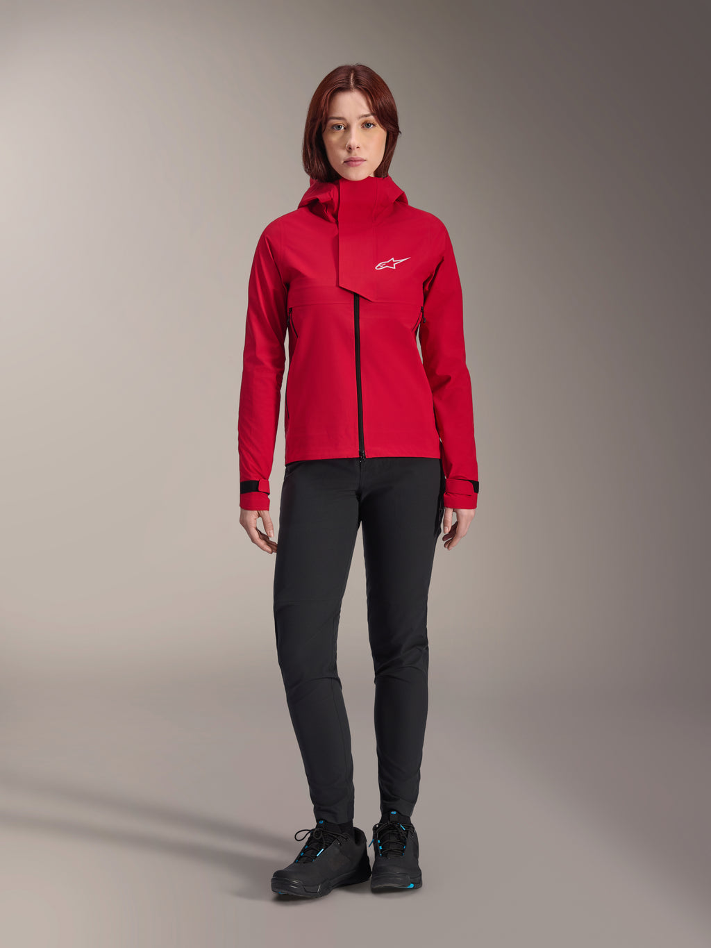 Alpinestars Stella A-Dura Elite Wasserdichte Jacke, Damen-MTB-Jacke, Red Fluo, getragen mit schwarzer Hose und Schuhen, mit hochgeschlossener Kapuze und weißem Alpinestars-Logo, technische Mountainbike-Bekleidung