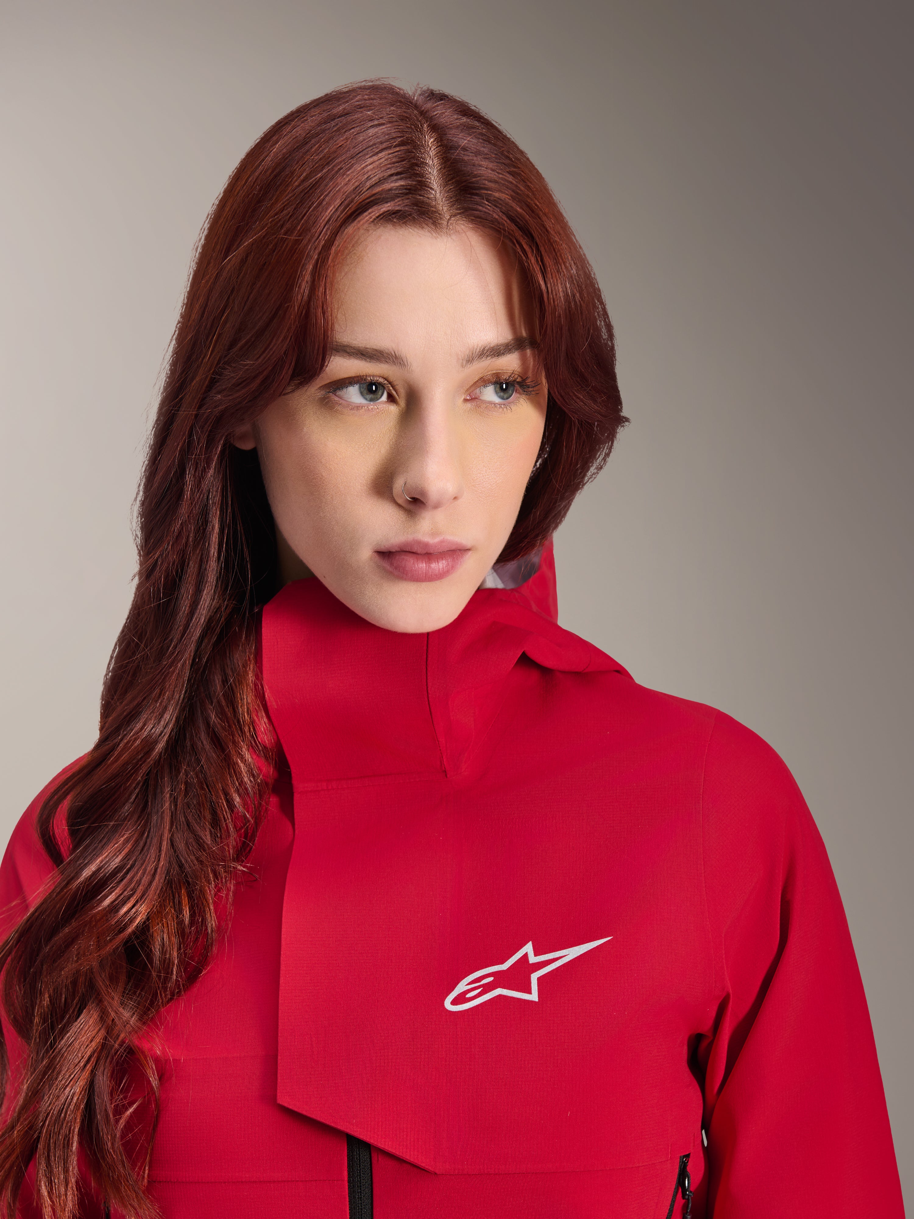 Alpinestars Stella A-Dura Elite Wasserdichte Jacke, Red Fluo, getragen von einer Frau mit langem rotem Haar, technische 3-lagige wasserdichte Jacke mit hohem Kragen und weißem Logo, Performance-Outdoor- und Mountainbike-Ausrüstung