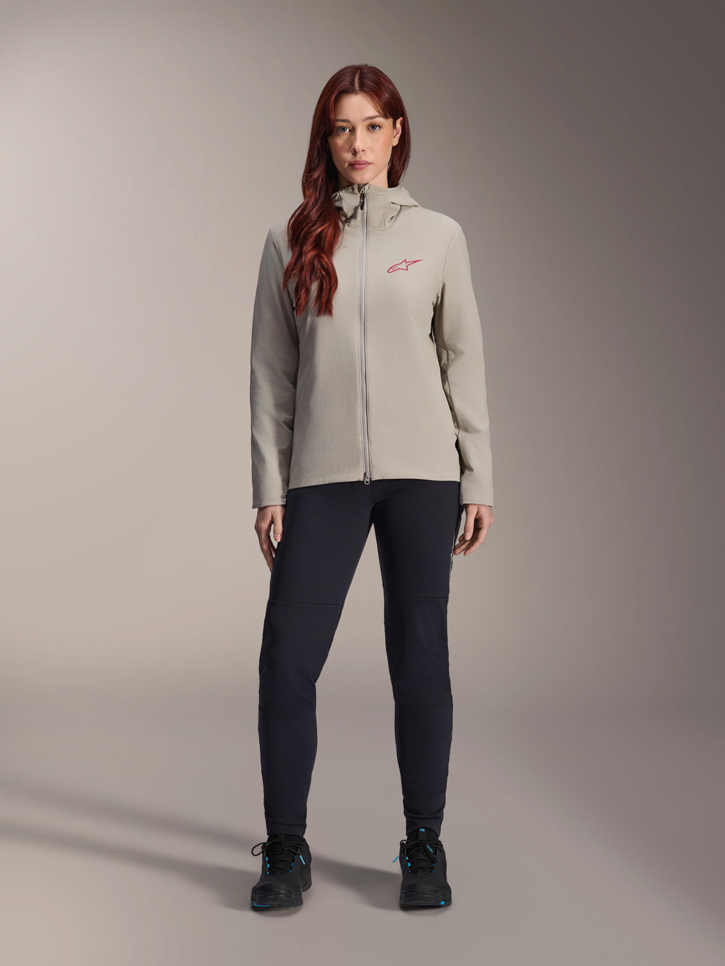 Alpinestars Stella A-Dura Thermal Damenjacke, Mountainbike-Jacke, Titanium-Grau, getragen mit schwarzer Funktionshose und schwarzen Schuhen, mit fleecegefütterter Kapuze und rotem Logo auf der Brust, konzipiert für Fahrten bei kaltem Wetter