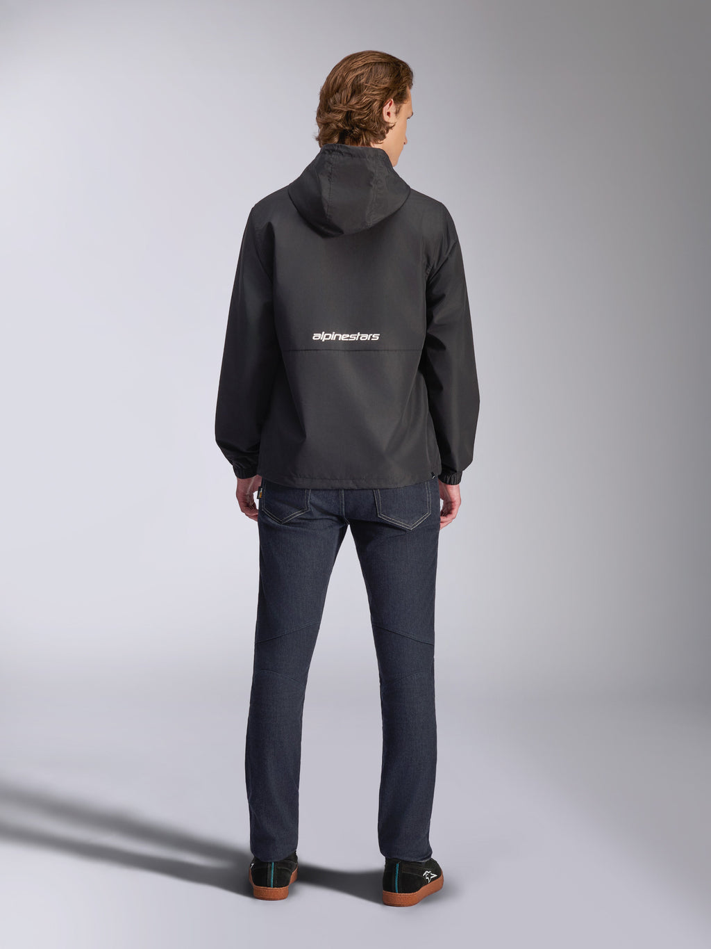 Alpinestars Speeder Chromium Windbreaker-Jacke, Schwarz, Rückansicht mit weißem Alpinestars-Branding in der Mitte, Modell mit Kapuze, getragen von einem Model mit dunkler Jeanshose, lässige, vom Motorsport inspirierte Jacke