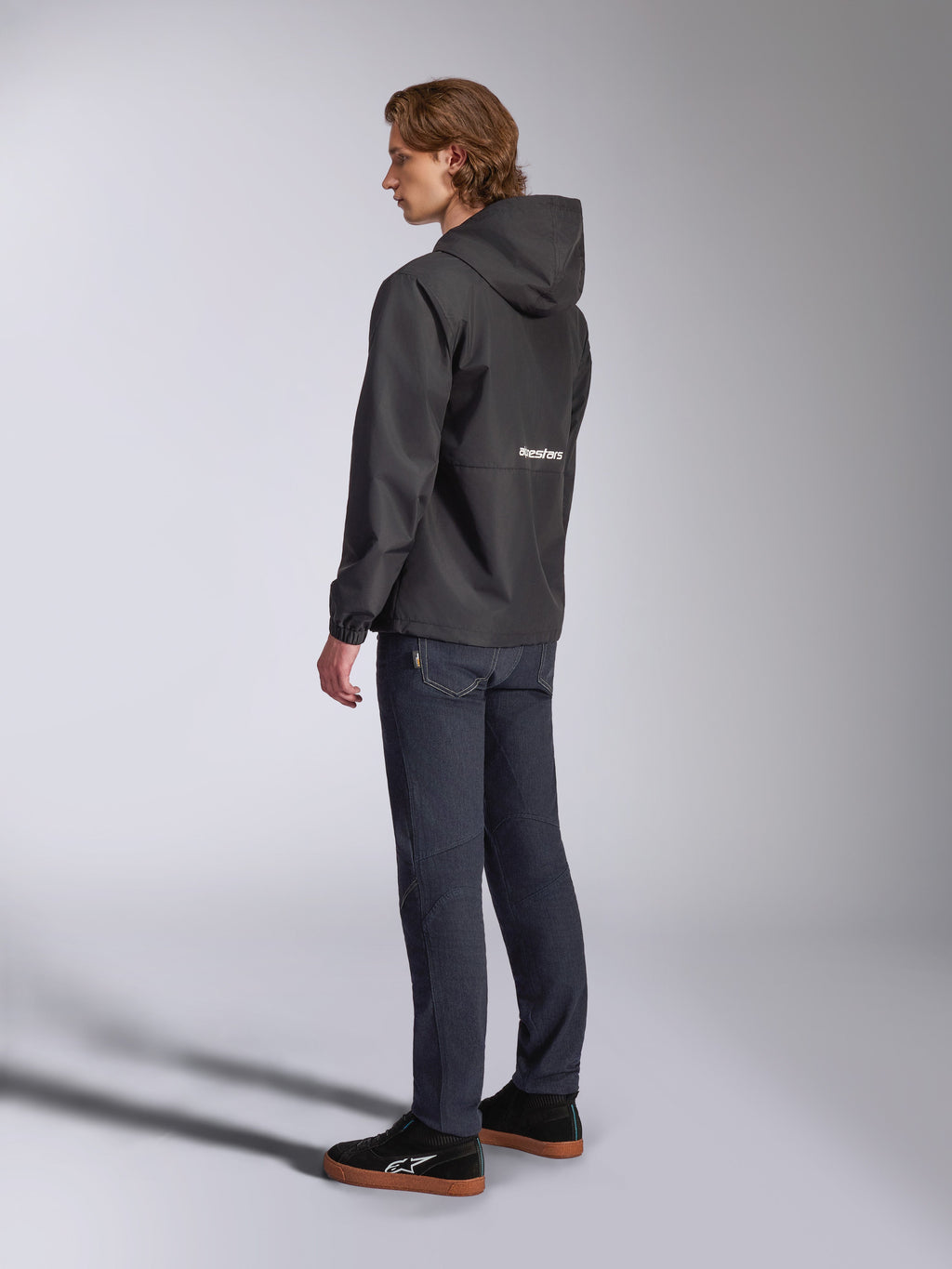 Alpinestars Speeder Chromium Windbreaker-Jacke, Schwarz mit weißem Logo auf der Rückseite, Herren-Freizeit-Windjacke mit Kapuze, Seitenansicht getragen mit dunkler Jeans und schwarzen Sneakern, Motorsport-inspirierte Streetwear