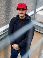 Man wearing the Alpinestars Overhaul Racer Jacket in Black, a hooded water-resistant rain shell jacket with a small chest logo, accessorized with a red Alpinestars cap, casual motorsport lifestyle apparel