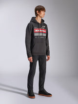 Alpinestars Harken Hoodie, schwarzer Fleece-Kapuzenpullover, große Brustgrafik mit roter Alpinestars Wortmarke und Stern-Logo, Kängurutasche, getragen von einem Model mit schwarzer Hose und Sneakern, lässige Motorsport-Streetwear