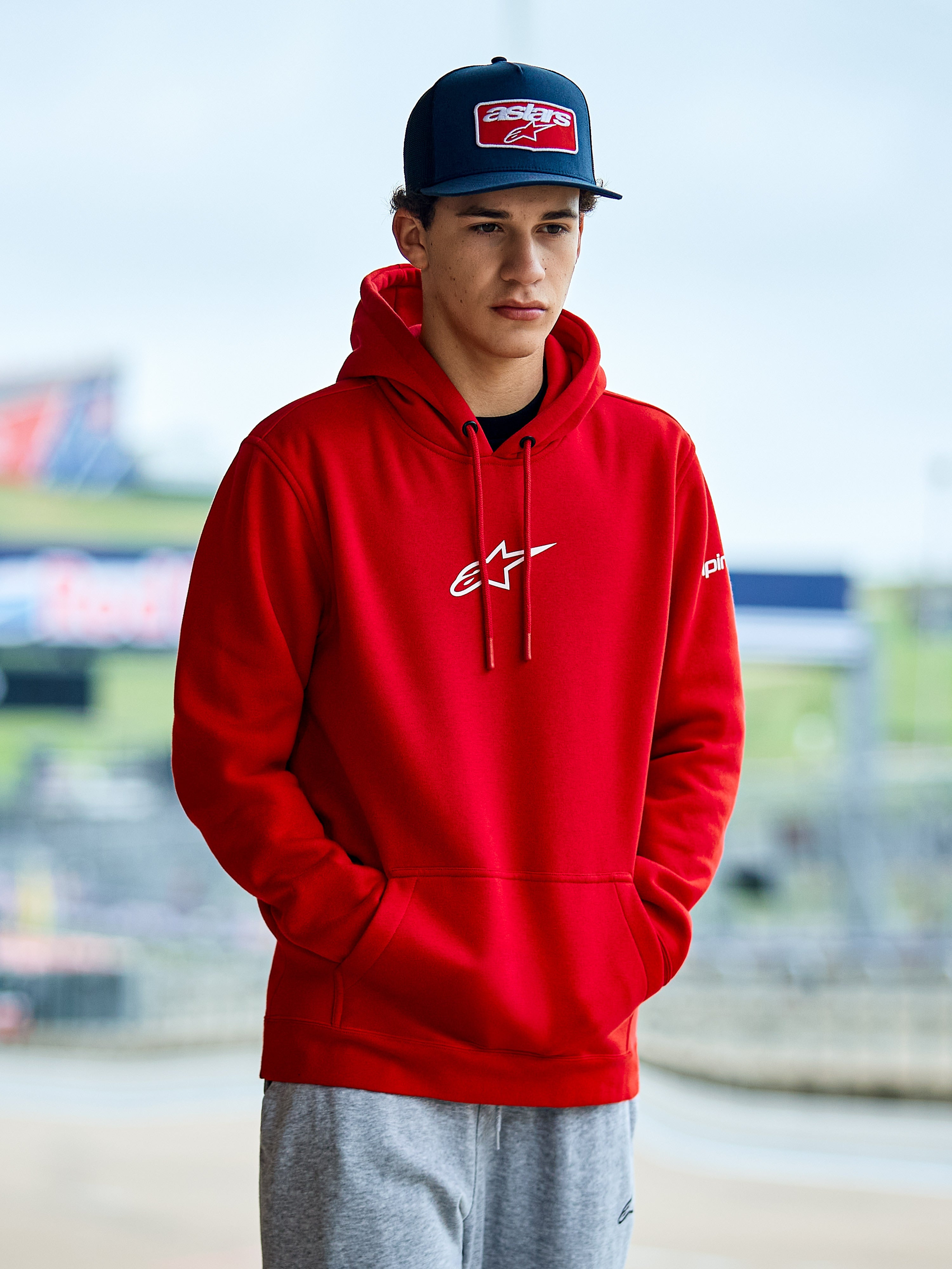 Alpinestars Frontal Hoodie, roter Fleece-Kapuzenpullover mit weißem Astars-Logo auf der Brust, Kängurutasche und Kapuze mit Kordelzug, getragen von einem Mann mit blauer Kappe, lässige Motorsport-Streetwear