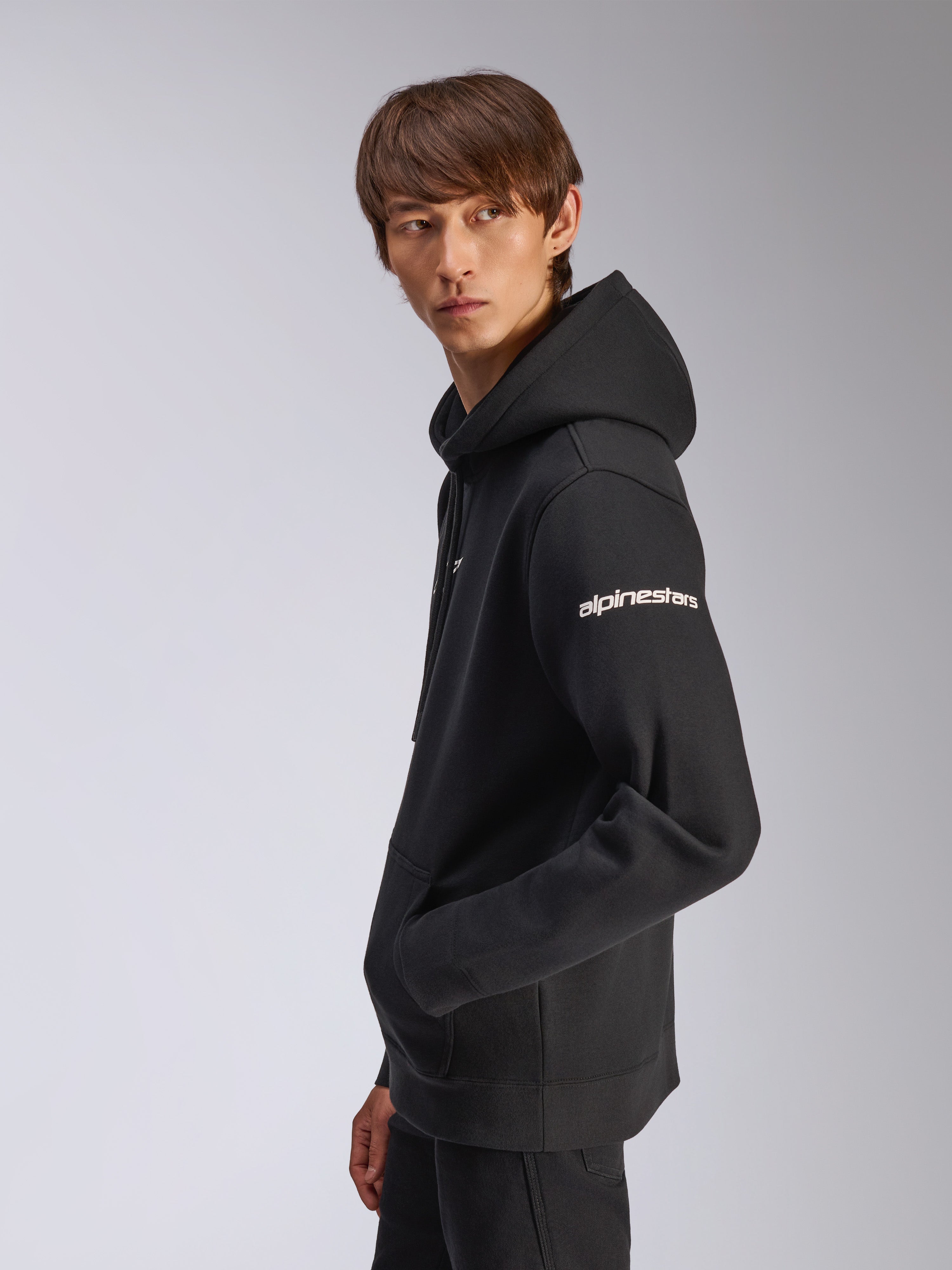 Alpinestars Frontal Hoodie, schwarzer Fleece-Pullover, Modell in Seitenansicht, weißer Alpinestars-Schriftzug auf dem Ärmel, Kängurutasche, lässige, vom Motorsport inspirierte Streetwear