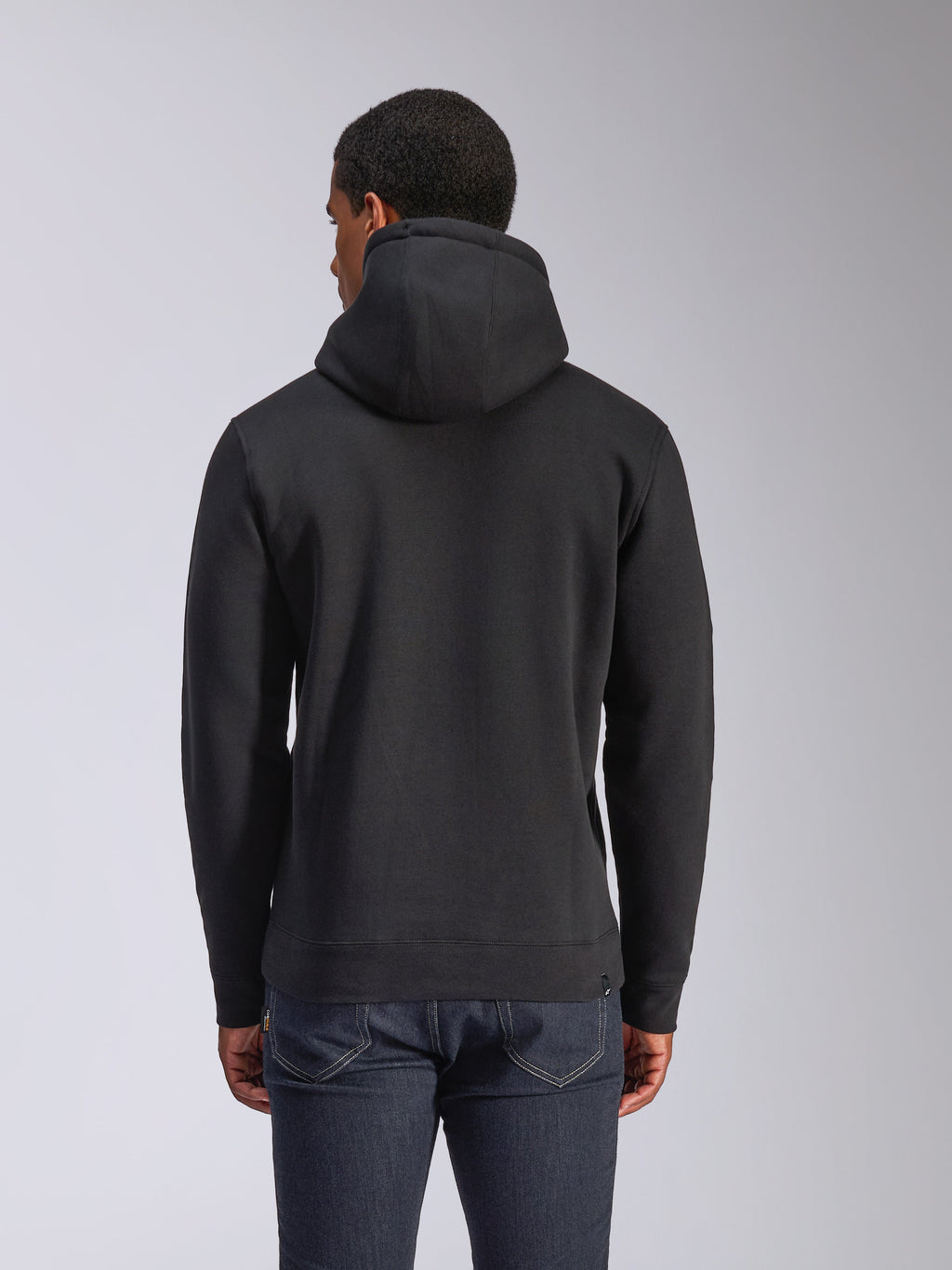 Alpinestars Blaze V3 Kapuzenpullover, Rückansicht eines schwarzen Fleece-Kapuzenpullovers, lässige Streetwear, mit Rippbündchen an Ärmeln und Saum, Kapuze mit Jersey-Futter, getragen mit dunkelblauen Jeans