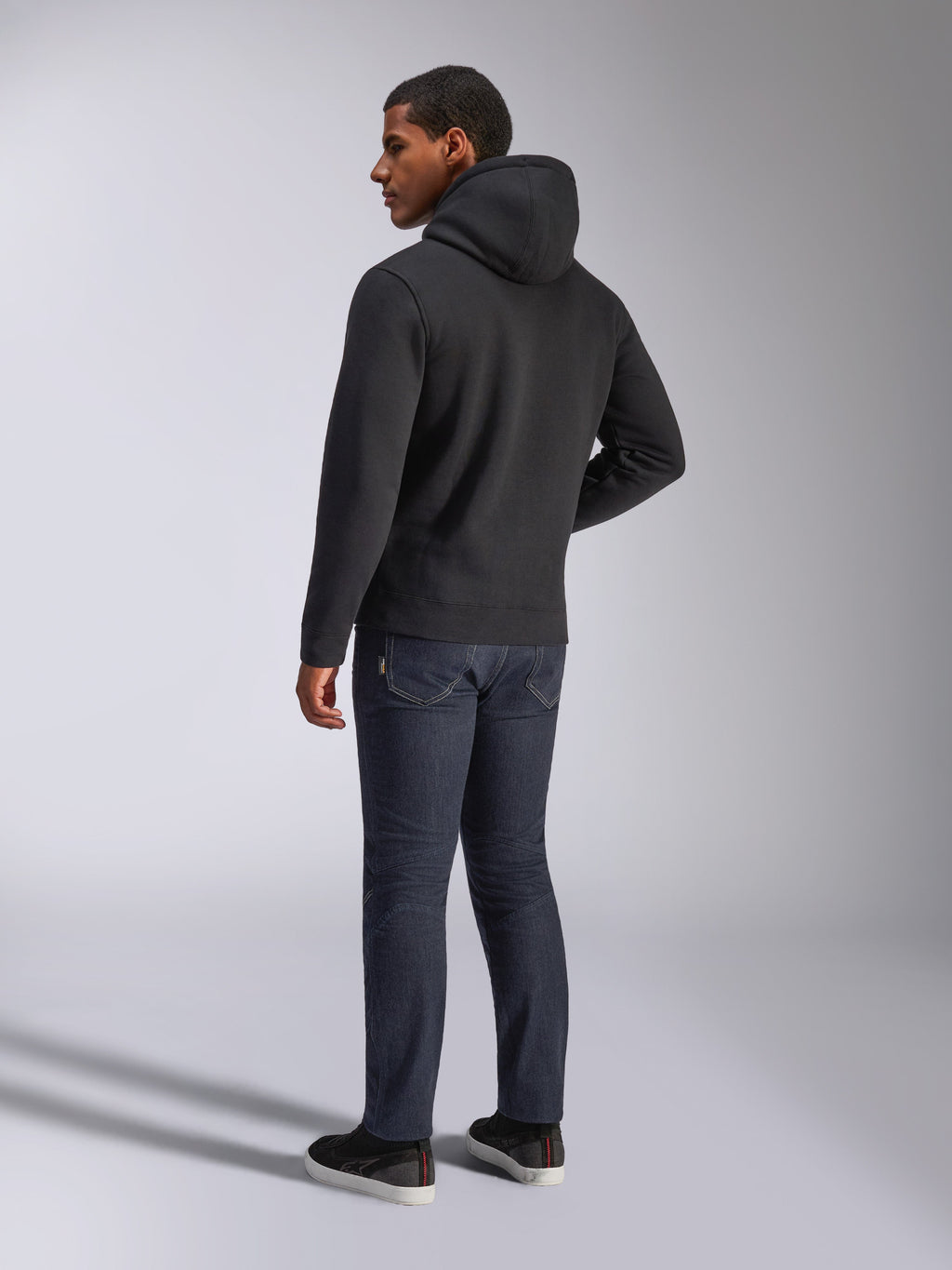 Alpinestars Blaze V3 Hoodie, Rückansicht eines Mannes im schwarzen Kapuzenpullover, kombiniert mit dunkler Jeans und schwarzen Sneakern, lässige Motorsport-inspirierte Streetwear