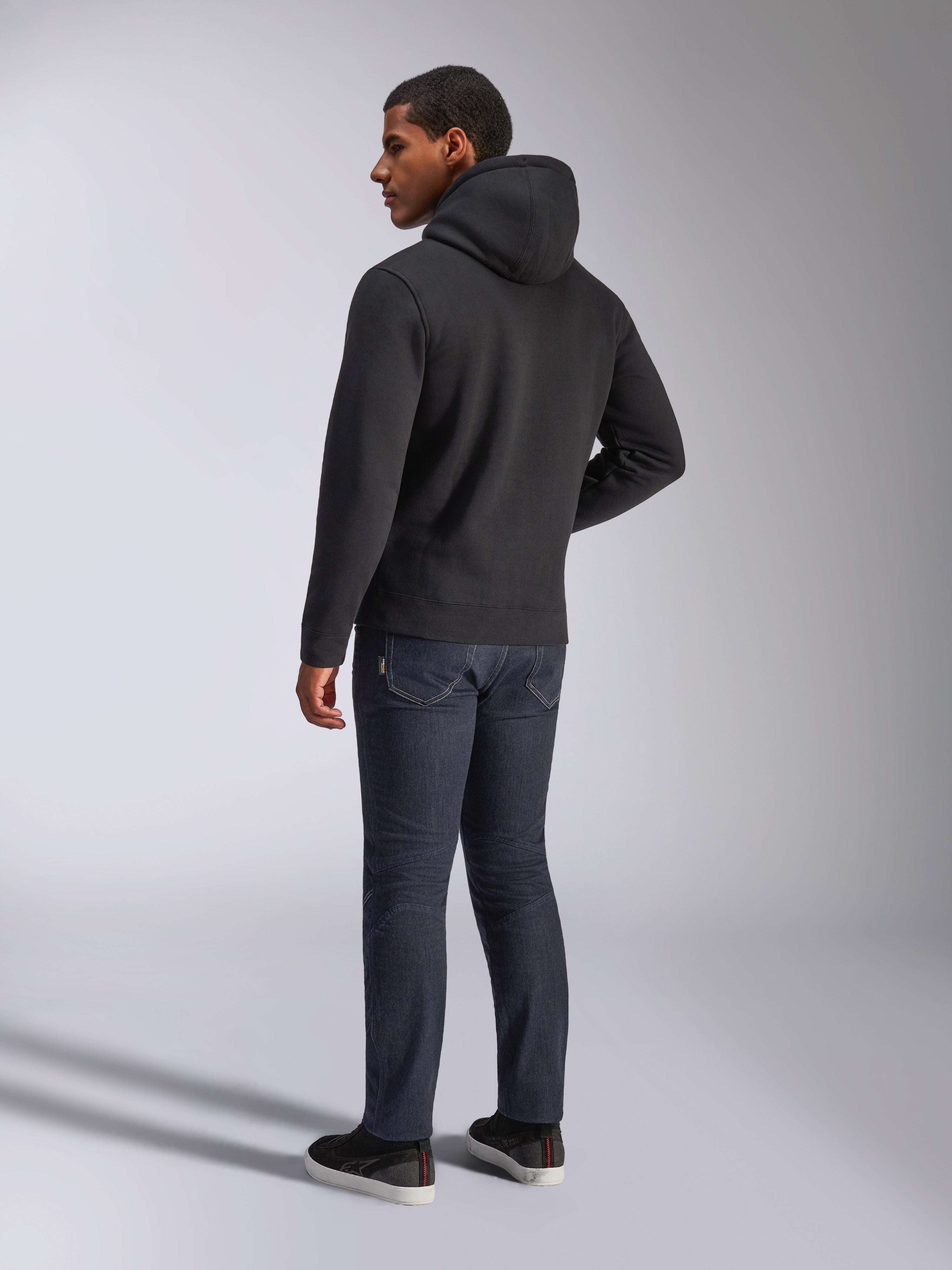 Alpinestars Blaze V3 Hoodie, Rückansicht eines Mannes im schwarzen Kapuzenpullover, kombiniert mit dunkler Jeans und schwarzen Sneakern, lässige Motorsport-inspirierte Streetwear
