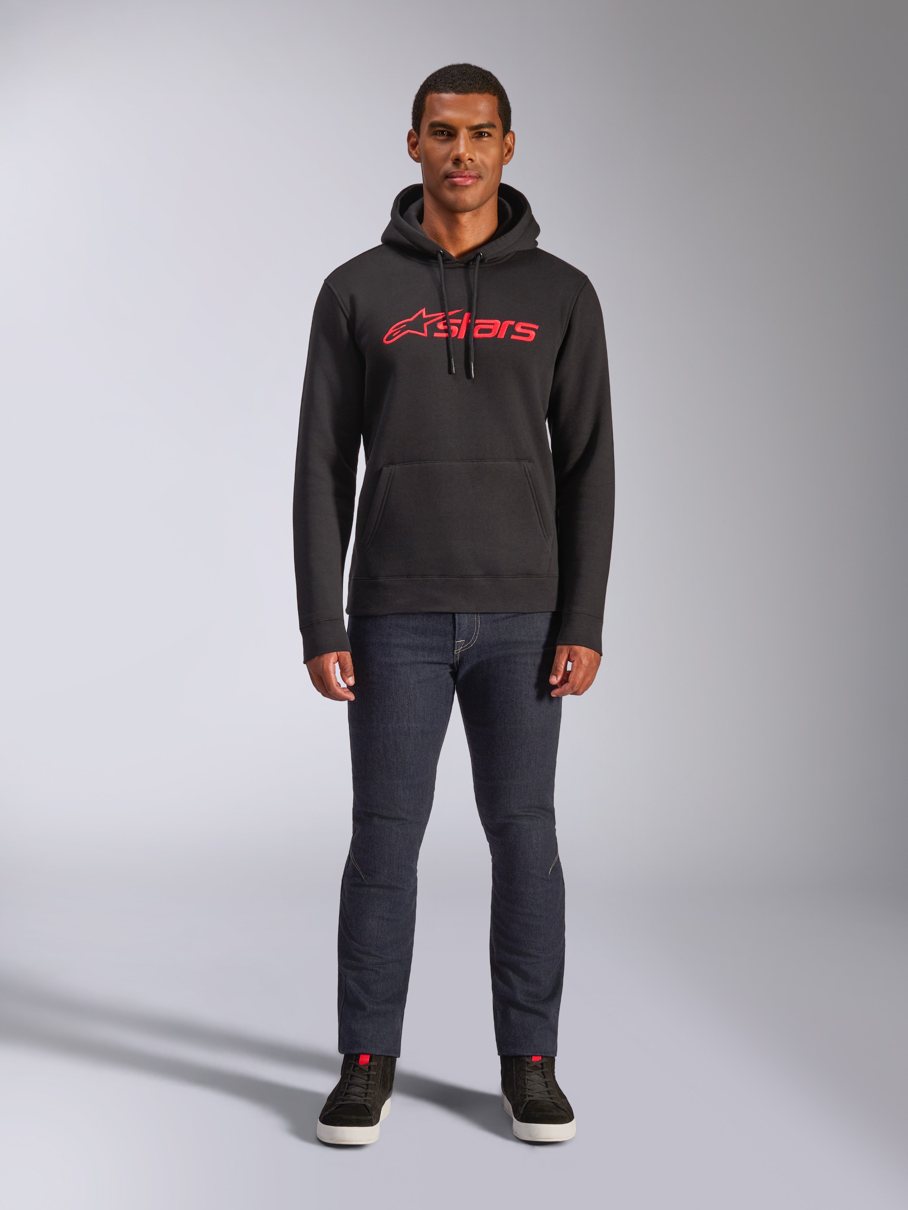 Alpinestars Blaze V3 Hoodie, lässige Streetwear, Schwarz mit rotem Astars-Logo, Vorderansicht, getragen von einem Model mit dunkler Jeanshose und schwarzen Sneakern, modernes, vom Motorsport inspiriertes Fleece-Design