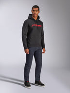 Alpinestars Blaze V3 Hoodie, schwarzer Fleece-Kapuzenpullover mit rotem Alpinestars-Logo auf der Brust, getragen von einem männlichen Model mit dunklen Denim-Jeans und schwarzen Sneakern, lässige Streetwear im Motorsport-Stil
