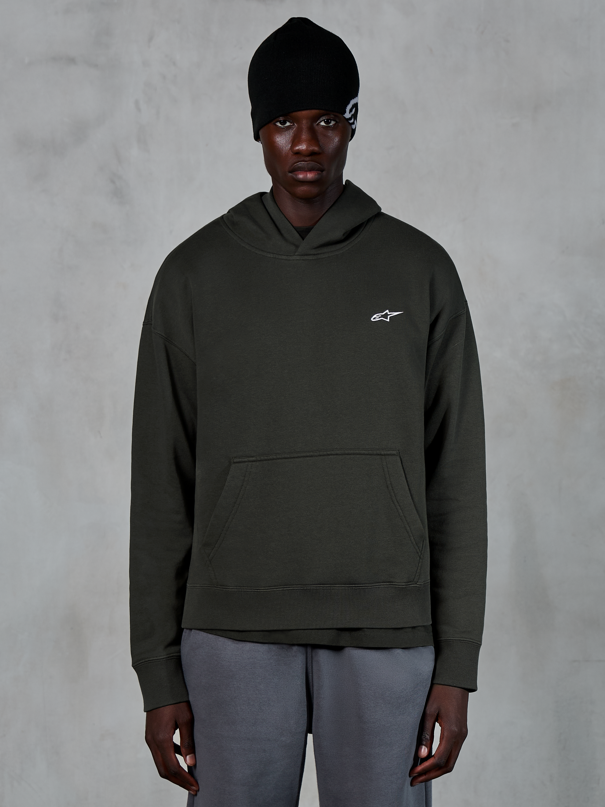 Alpinestars Essentials Ageless Hoodie, lässige Streetwear, Off-Black-Pullover mit einem kleinen weißen Alpinestars-Logo auf der Brust, getragen mit einer schwarzen Beanie, minimalistisches Design mit Kängurutasche vorne und Stone-Wash-Finish