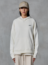 Alpinestars Essentials Ageless Hoodie, off-white pullover fleece hoodie, female model wearing tan cap and matching cream joggers, small black Alpinestars logo on chest, front kangaroo pocket, relaxed fit, casual motorsport-inspired streetwear