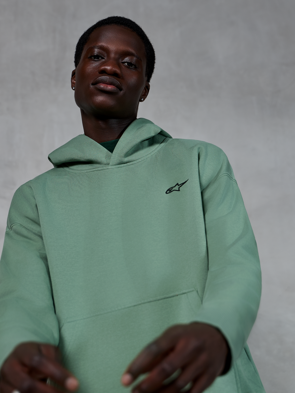 Alpinestars Essentials Ageless Hoodie, lässige Sportbekleidung in Cactus Grün, getragen von einem Model, mit kleinem schwarzen Alpinestars-Logo auf der Brust, hochwertiges Fleece mit entspannter Passform und cleaner Ästhetik.