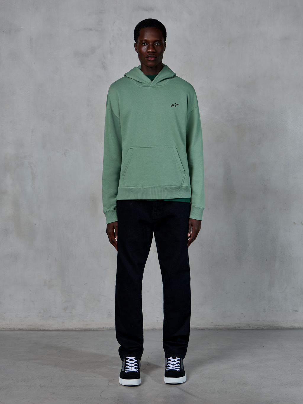 Alpinestars Essentials Ageless Hoodie, Kaktusgrün, kleines schwarzes Brustlogo, Kängurutasche, lässige Lifestyle-Streetwear getragen mit schwarzer Hose und schwarz-weißen Sneakern vor einer strukturierten Wand