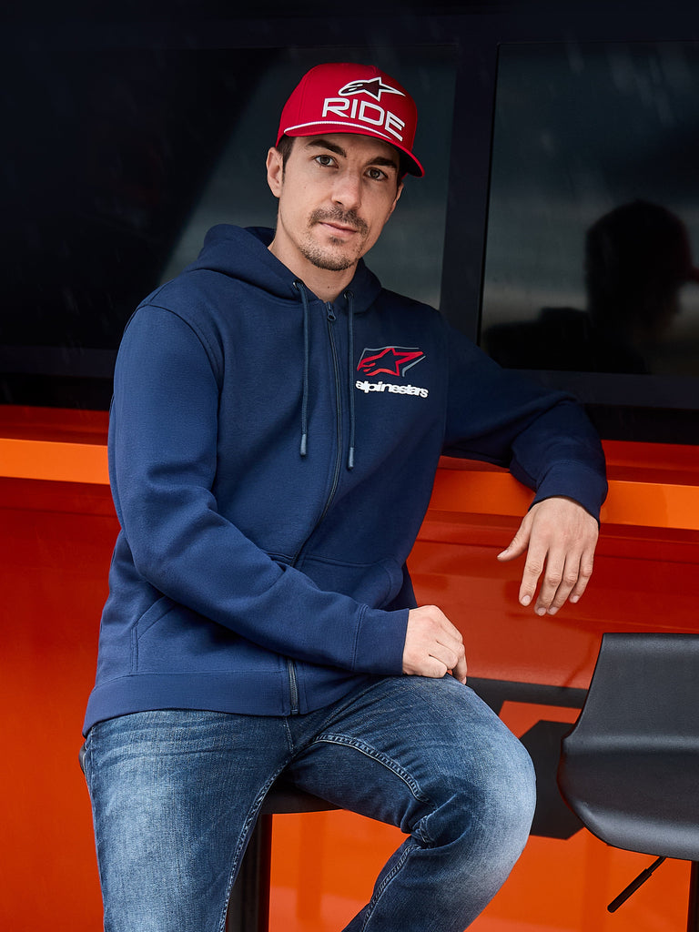 Alpinestars Formulation Hoodie, Dunkelblau, Kapuzenjacke mit Reißverschluss und Alpinestars-Logo auf der Brust, getragen mit einer roten Kappe und Jeans, lässige Motorsport-Lifestyle-Streetwear