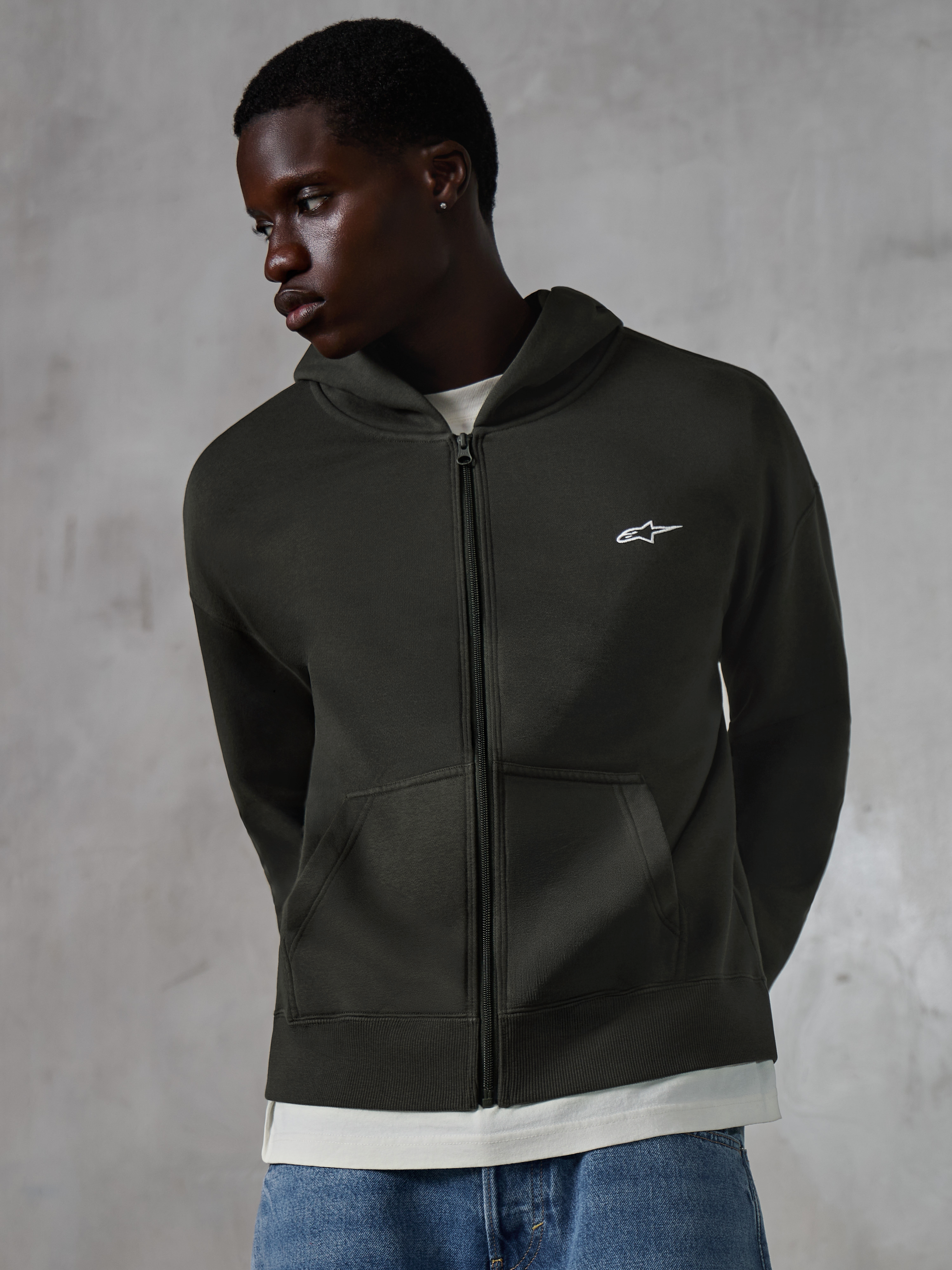 Alpinestars Essentials Ageless Zip Hoodie, Off-Black Streetwear, Fleece-Jacke mit durchgehendem Reißverschluss und kleinem weißen Alpinestars-Logo auf der Brust, getragen von einem Model mit blauen Jeans, entspannte Passform mit Vordertaschen und Stonewash-Finish.