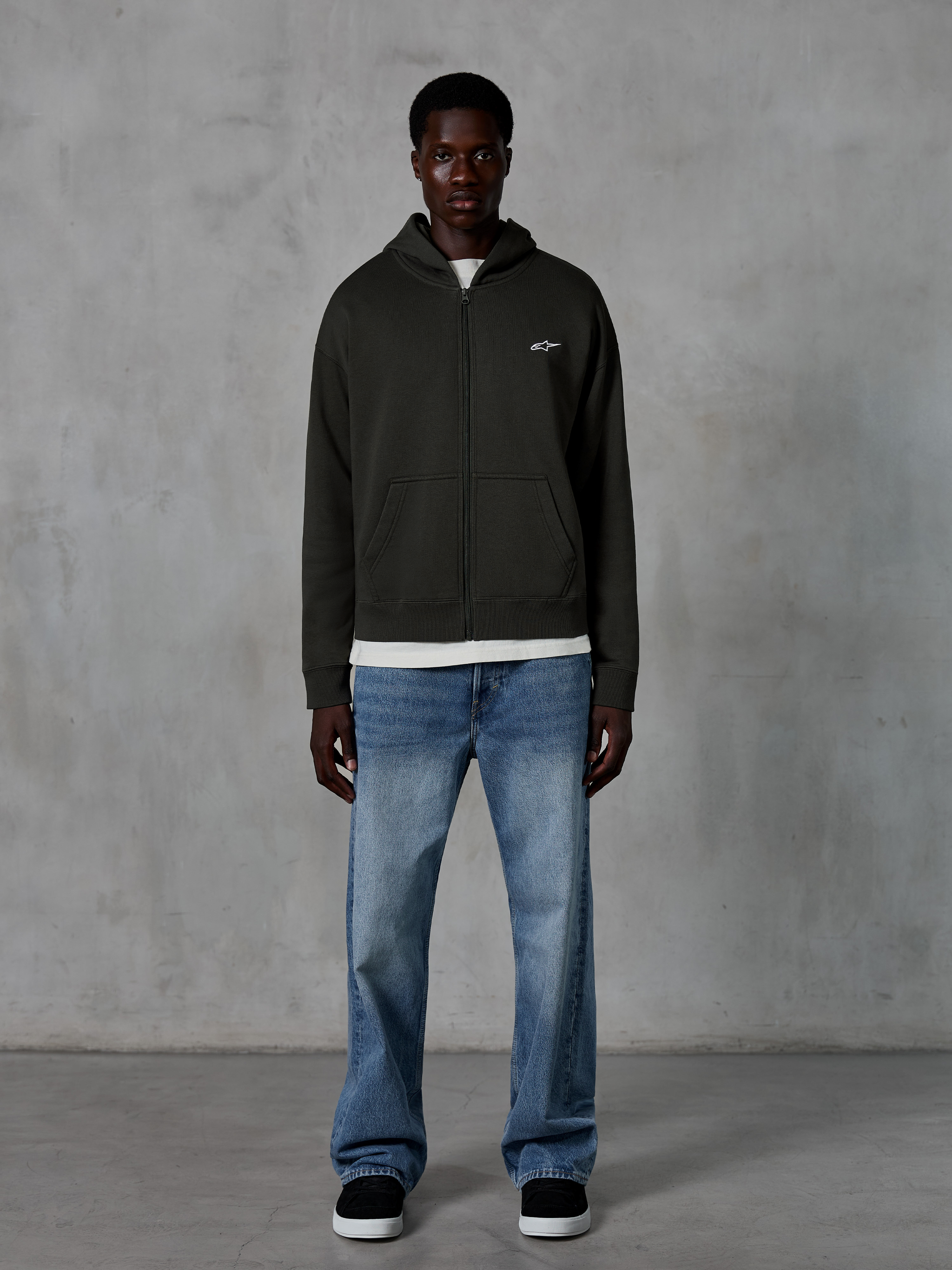 Alpinestars Essentials Ageless Zip Hoodie, Kapuzenjacke mit Reißverschluss, Off Black, mit einem kleinen weißen Alpinestars-Logo auf der Brust, getragen von einem Model mit weißem T-Shirt und blauen Jeans, entspannte Passform, lässige Motorsport-Streetwear