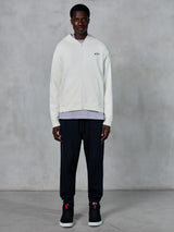 Alpinestars Essentials Ageless Zip Hoodie, cremeweißer Hoodie mit durchgehendem Reißverschluss, entspannte Passform mit kleinem schwarzem Logo auf der Brust, kombiniert mit schwarzer Jogginghose und Sneakern, lässige, vom Motorsport inspirierte Streetwear.