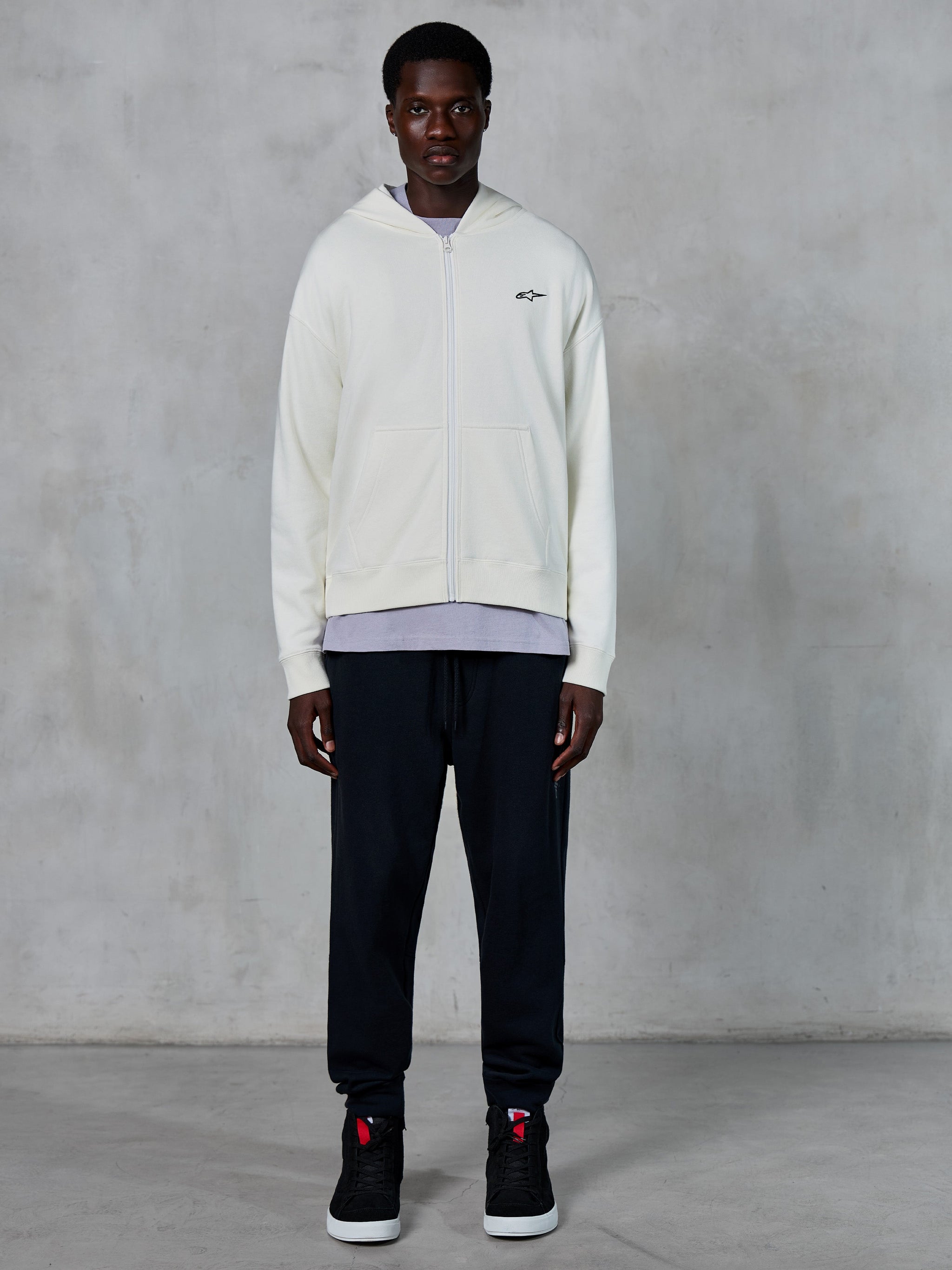 Alpinestars Essentials Ageless Zip Hoodie, cremeweißer Hoodie mit durchgehendem Reißverschluss, entspannte Passform mit kleinem schwarzem Logo auf der Brust, kombiniert mit schwarzer Jogginghose und Sneakern, lässige, vom Motorsport inspirierte Streetwear.