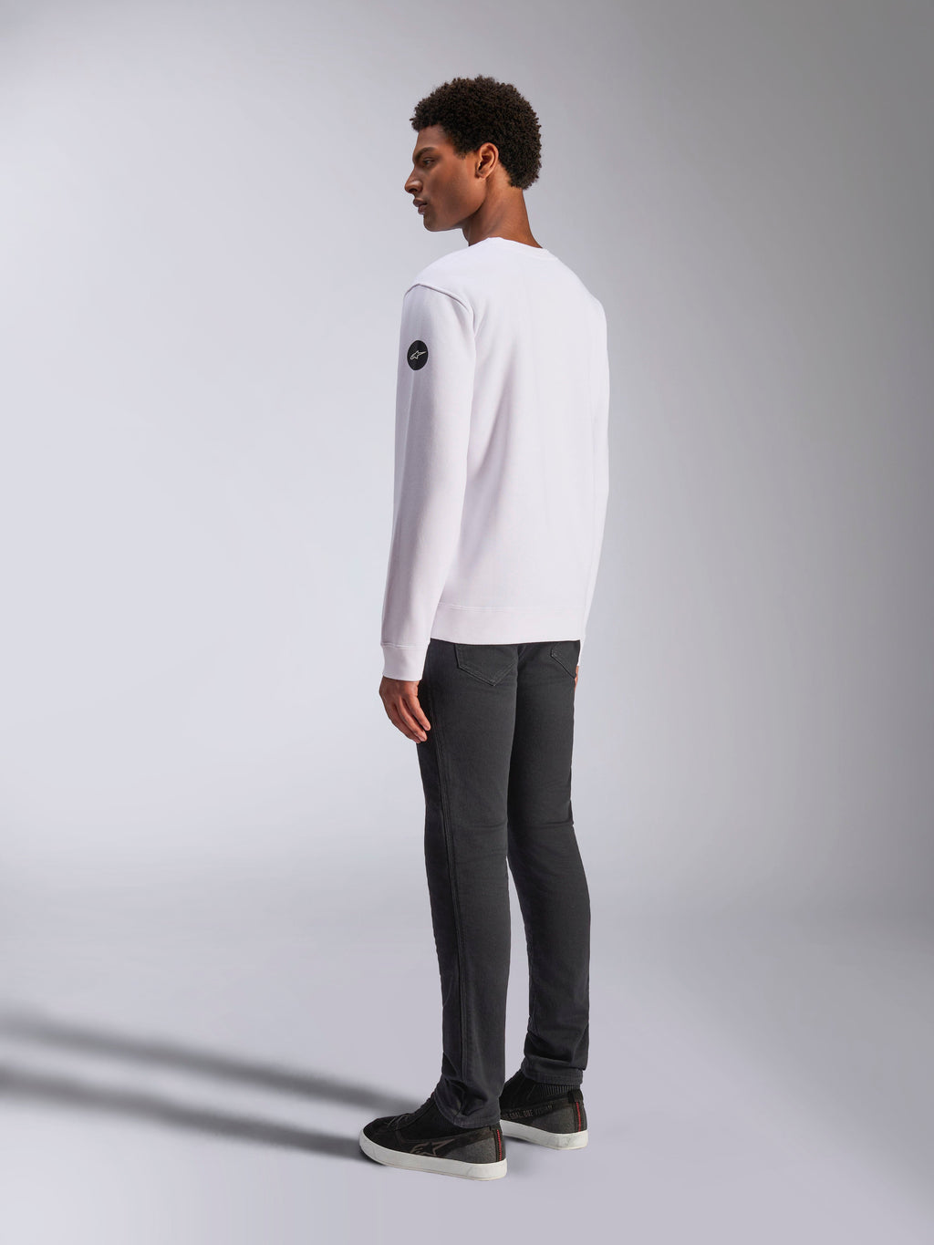 Alpinestars Bettern Crew, weißes Crewneck-Sweatshirt, Rück- und Seitenansicht, schwarzes kreisförmiges Logo am Ärmel, getragen mit dunkelgrauer Hose und schwarzen Sneakern, lässige Motorsport-Lifestyle-Streetwear