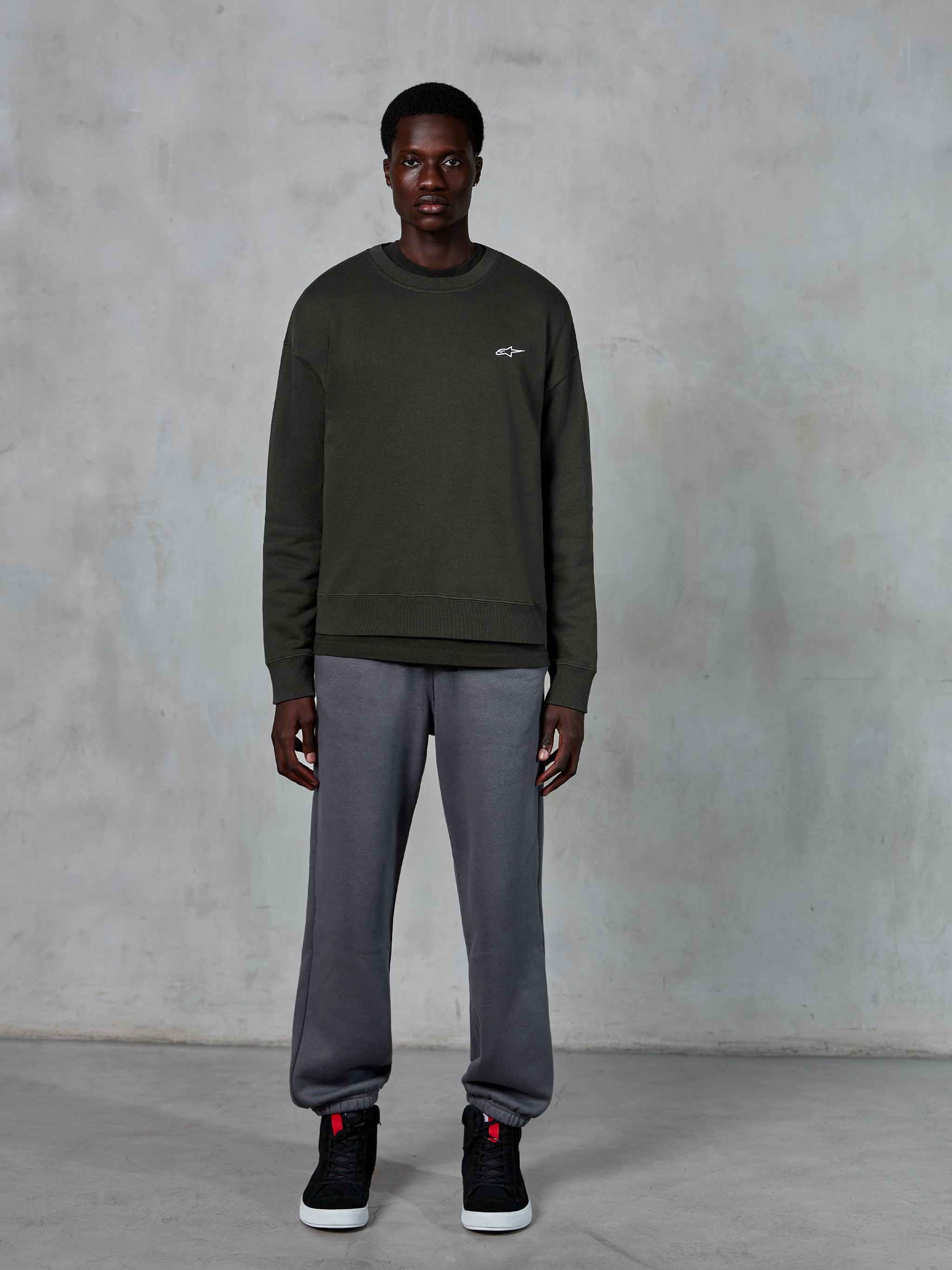 Alpinestars Essentials Ageless Crew, dark green crewneck sweatshirt with a small white logo on the chest, worn by a model with gray sweatpants and black high-top sneakers, casual lifestyle apparel.
