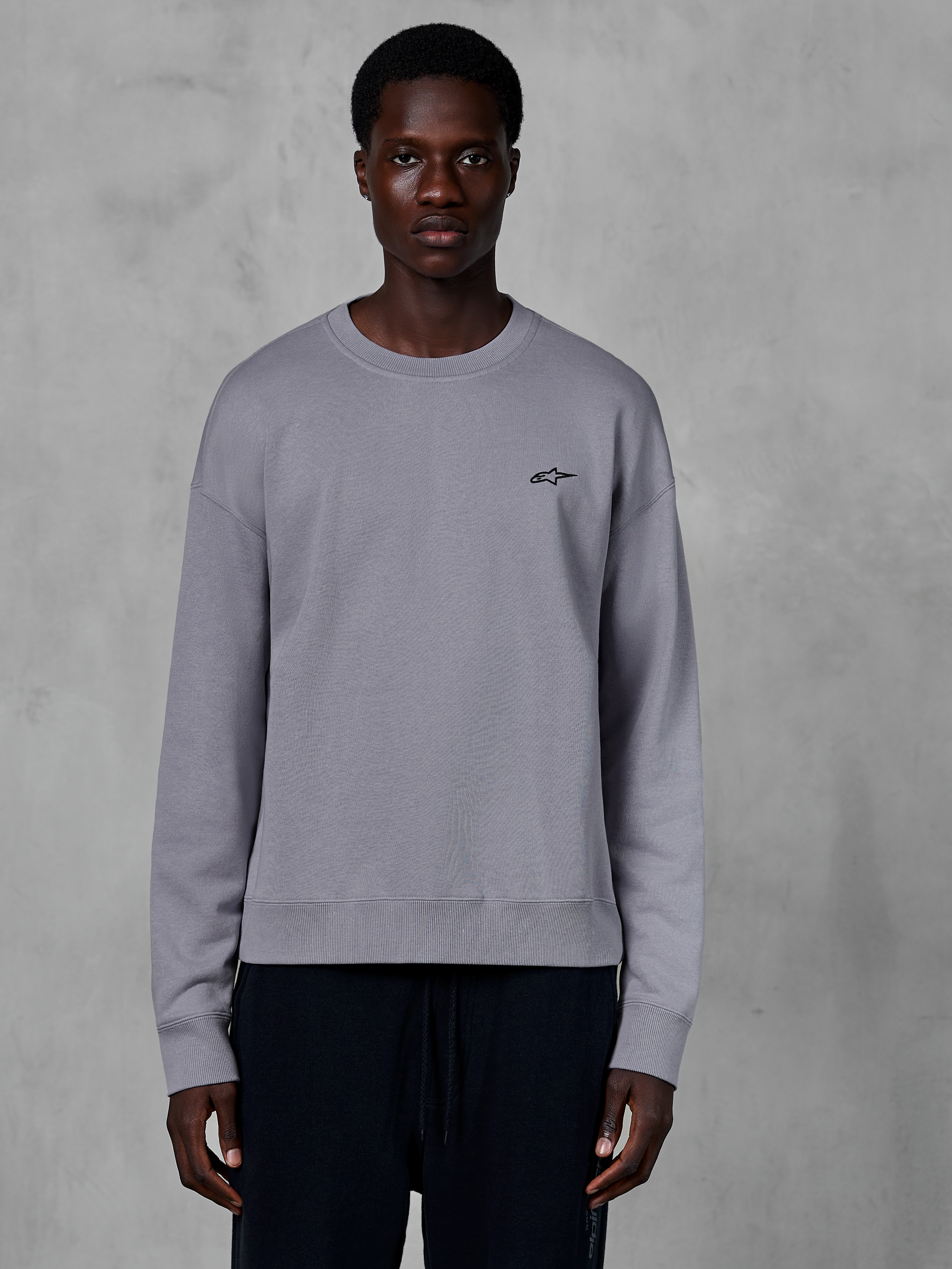 Alpinestars Essentials Ageless Crew, silbernes Rundhals-Sweatshirt von einem Model getragen, mit einem kleinen schwarzen Stern-Logo auf der Brust, entspannte Passform mit Rippbündchen und Saum, lässige Motorsport-Streetwear