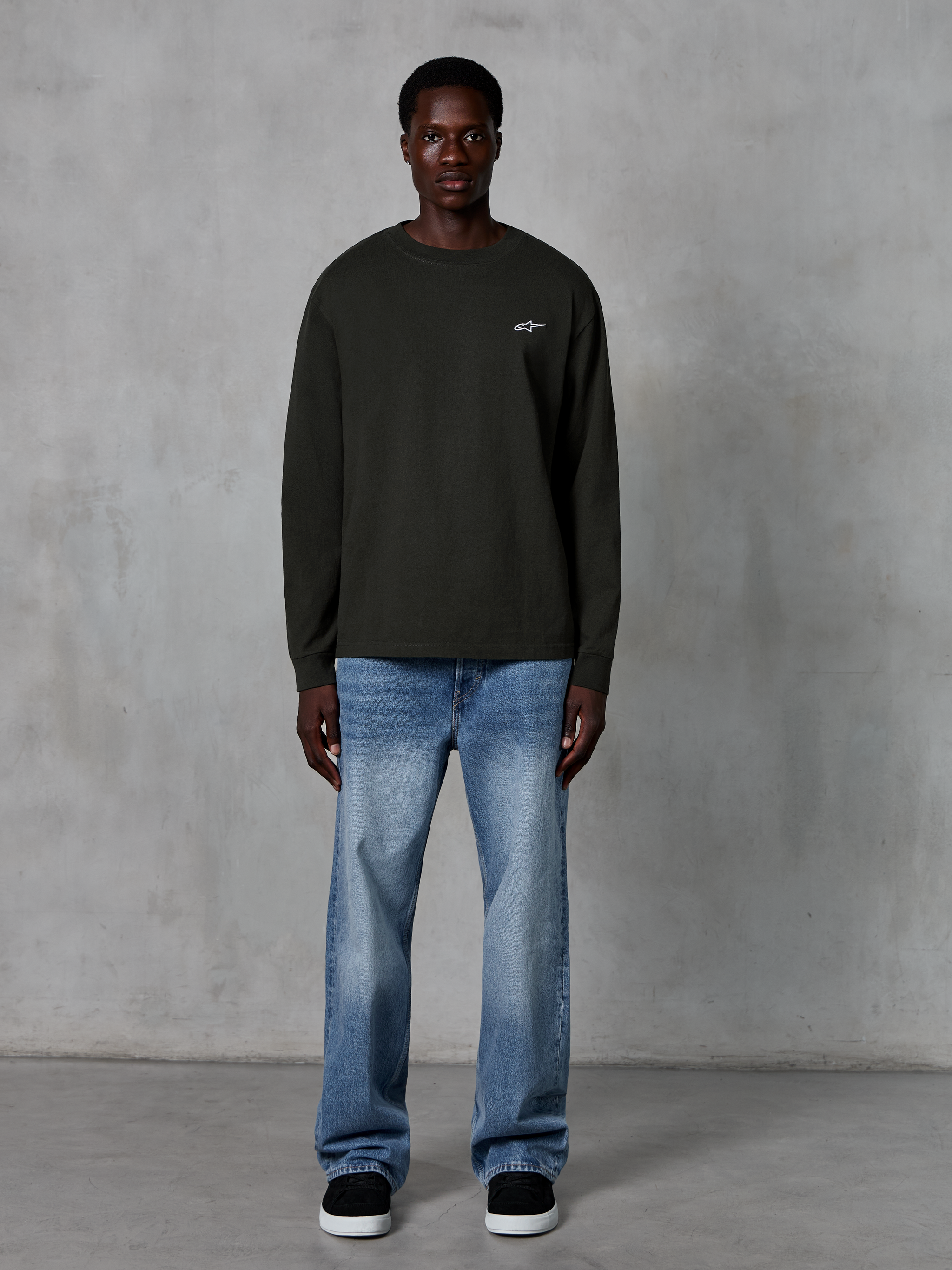 Alpinestars Essentials Ageless LS Tee, Langarm-T-Shirt, Off-Black, mit kleinem weißen Sternlogo auf der Brust, entspannte Passform, lässige Motorsport-Streetwear, kombiniert mit hellblauen Jeans