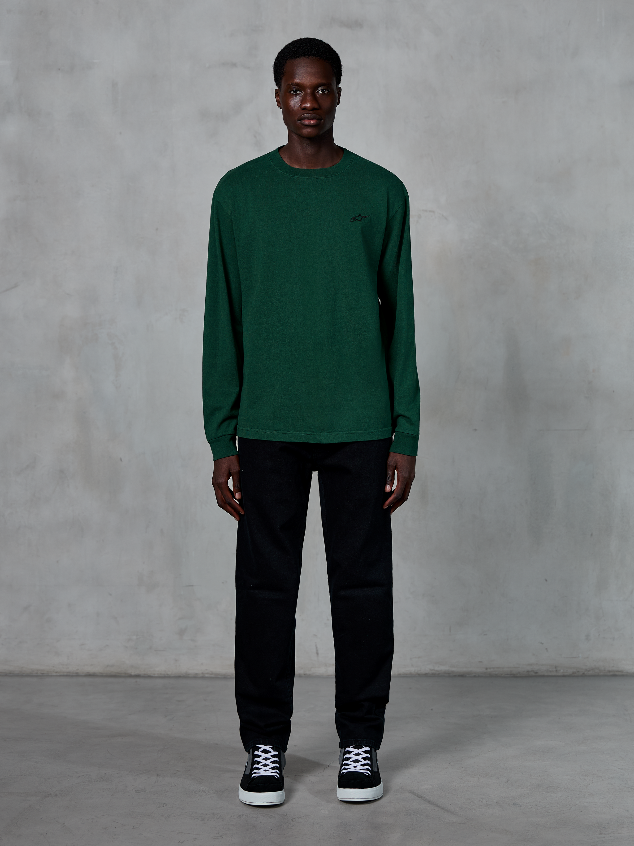 Alpinestars Essentials Ageless LS Tee, Langarm-T-Shirt, Tannengrün, mit kleinem schwarzen Logo auf der Brust, getragen von einem Model mit schwarzer Hose und Sneakern, lässiger Streetwear-Stil